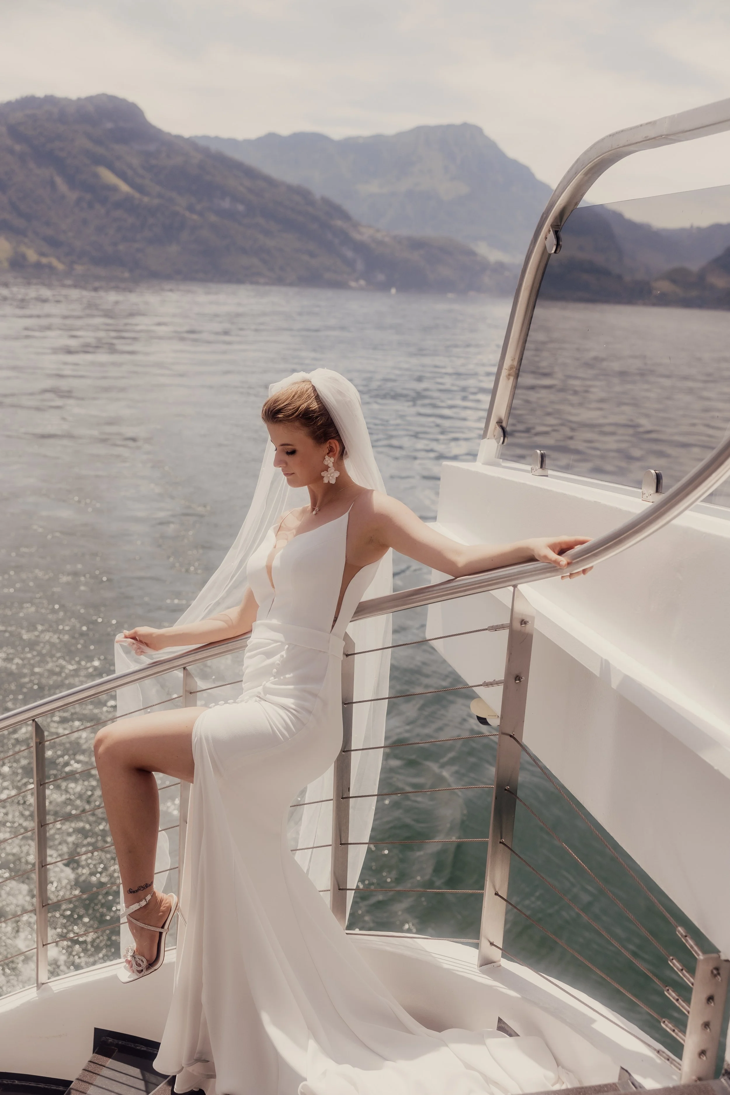Bride in a white dress and veil standing on a boat's railing, scenic mountains and lake in the background.