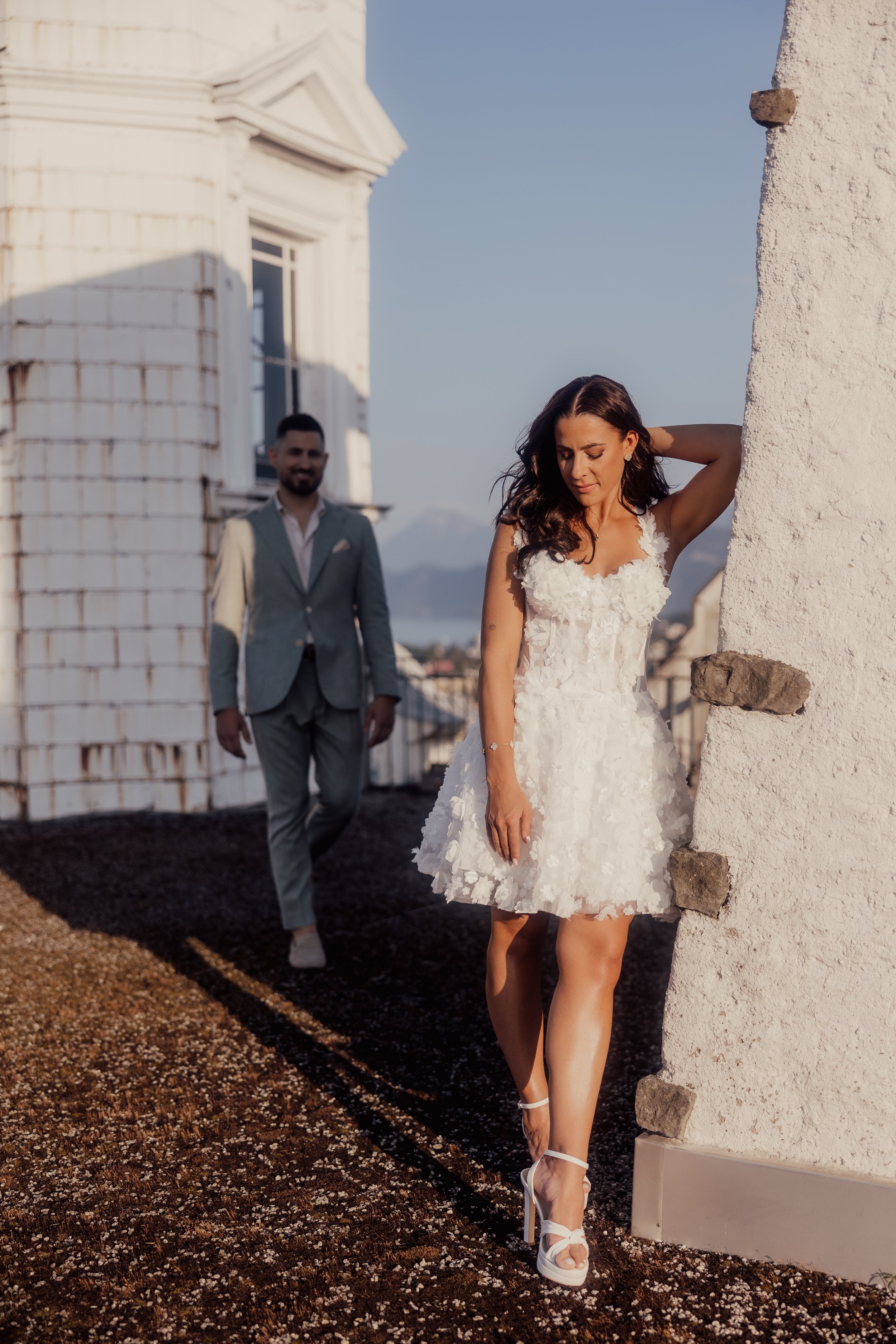 A woman in a white floral dress and high heels poses against a wall, with a man in the background wearing a suit. They appear to be on a rooftop with a view of mountains in the distance.