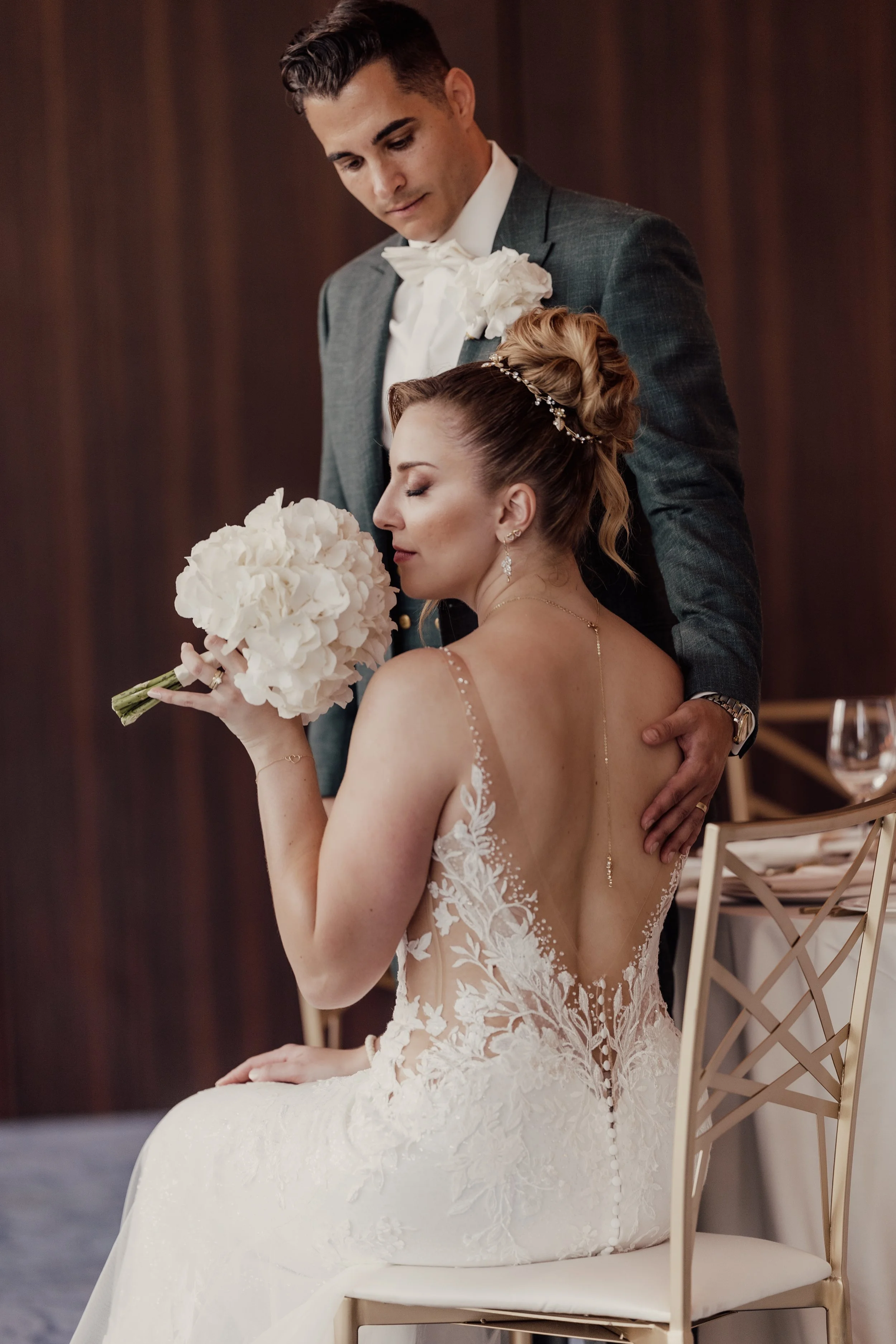 Bride and groom in elegant wedding attire; the bride is sitting and holding a bouquet of white flowers, while the groom stands behind her.