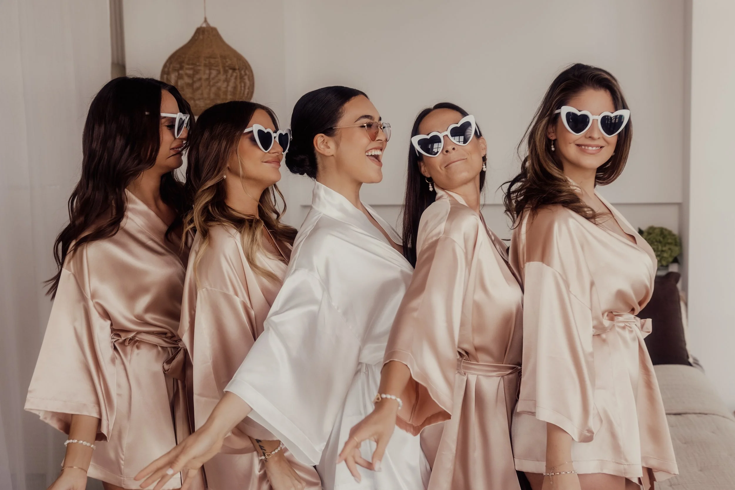 Group of women in silk robes and heart-shaped sunglasses laughing and posing indoors.