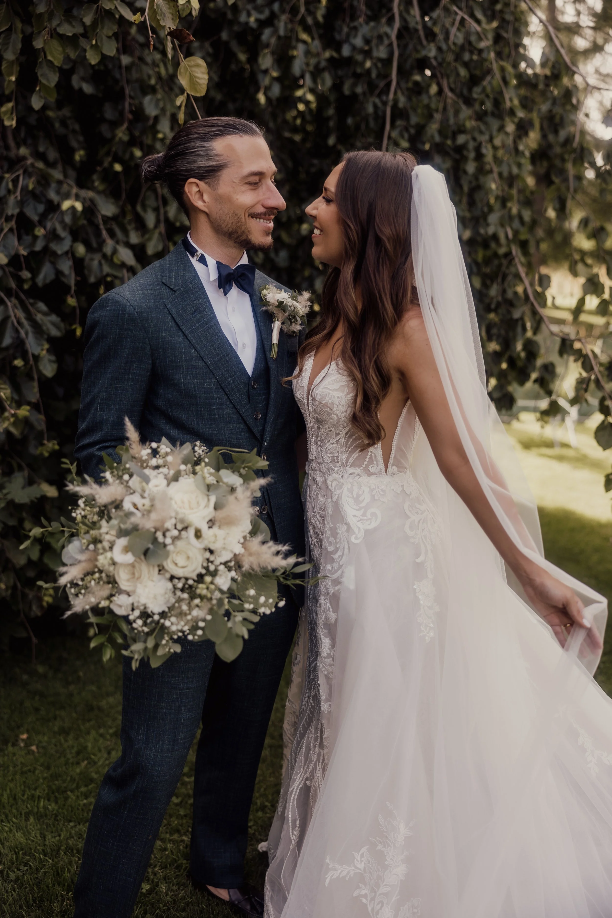 Bride and groom smiling at each other outdoors, bride in a white lace gown holding a bouquet, groom in a dark suit.