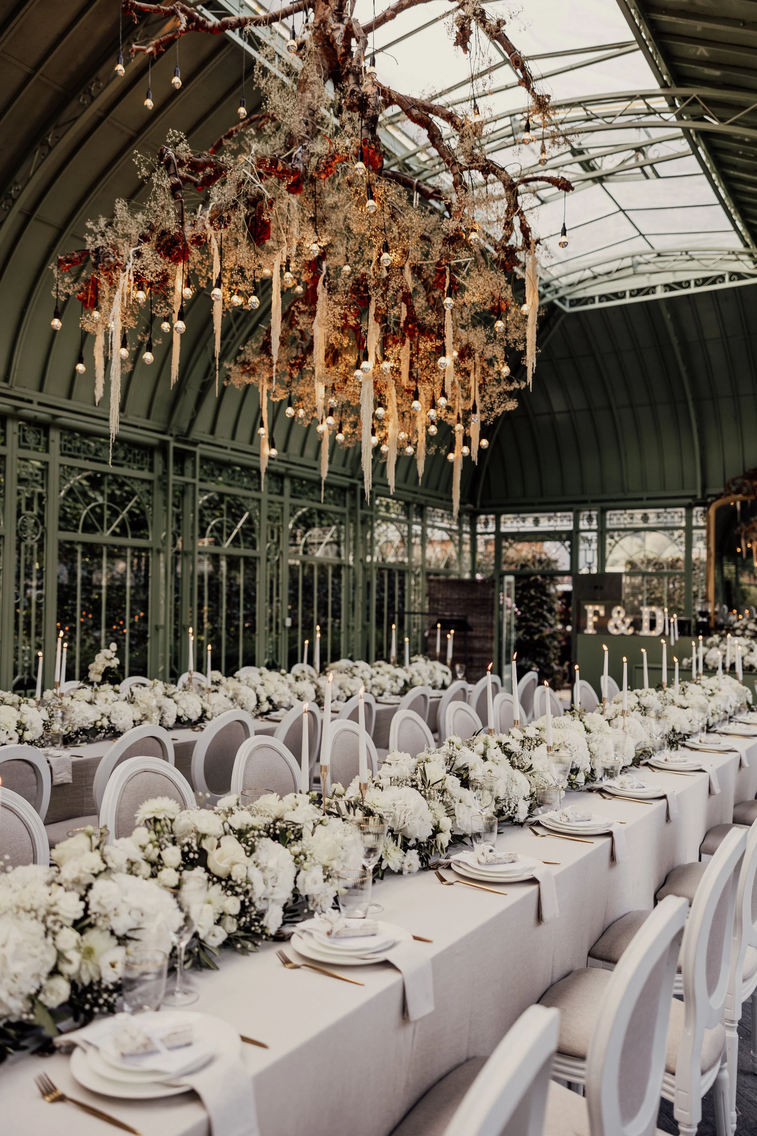 Elegant wedding reception setup with long tables adorned with white floral arrangements and candles, under an ornate chandelier with hanging greenery and lights in a greenhouse setting.