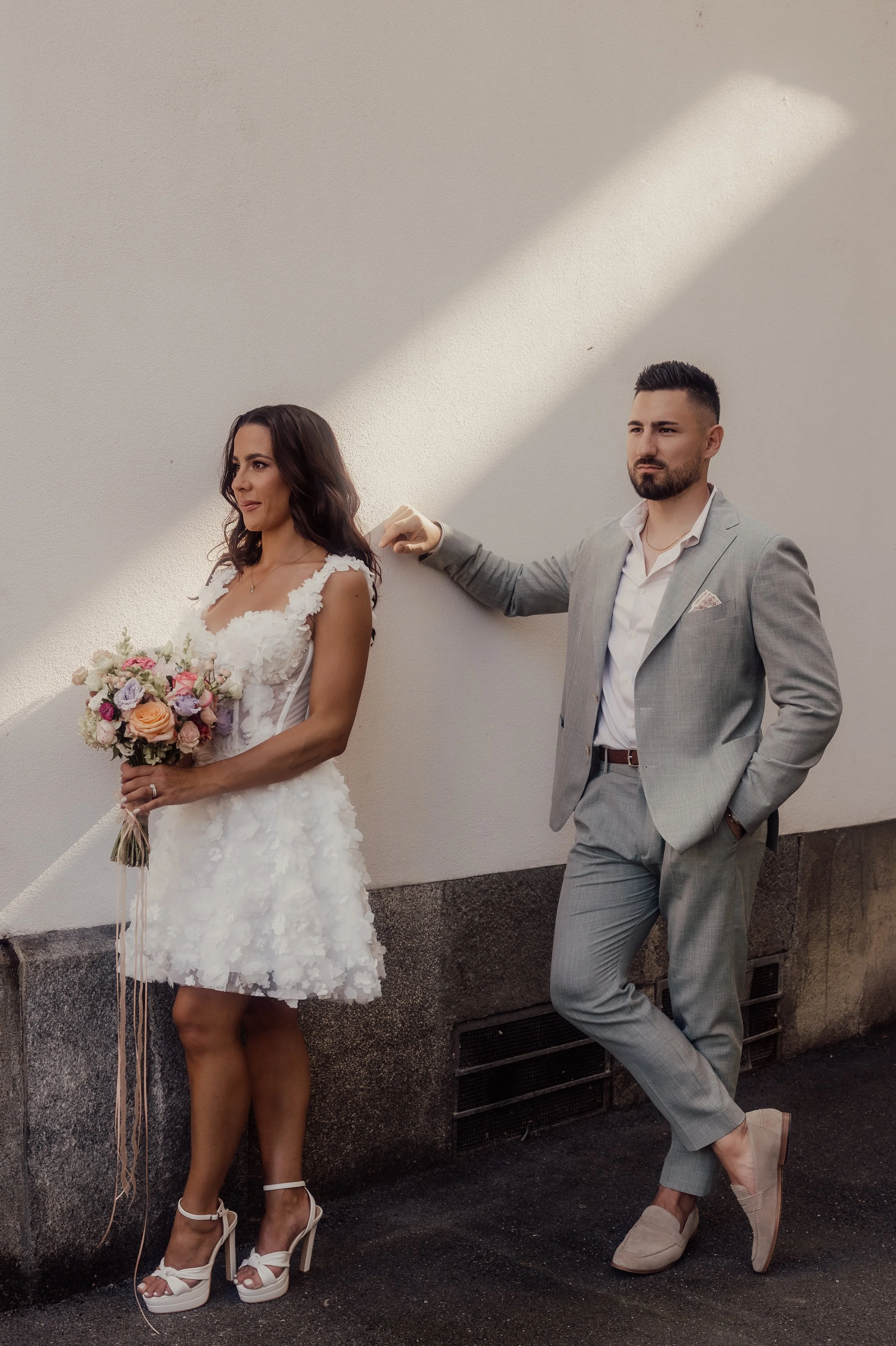A woman in a short white wedding dress holding a bouquet stands next to a man in a gray suit, both posed against a white wall.