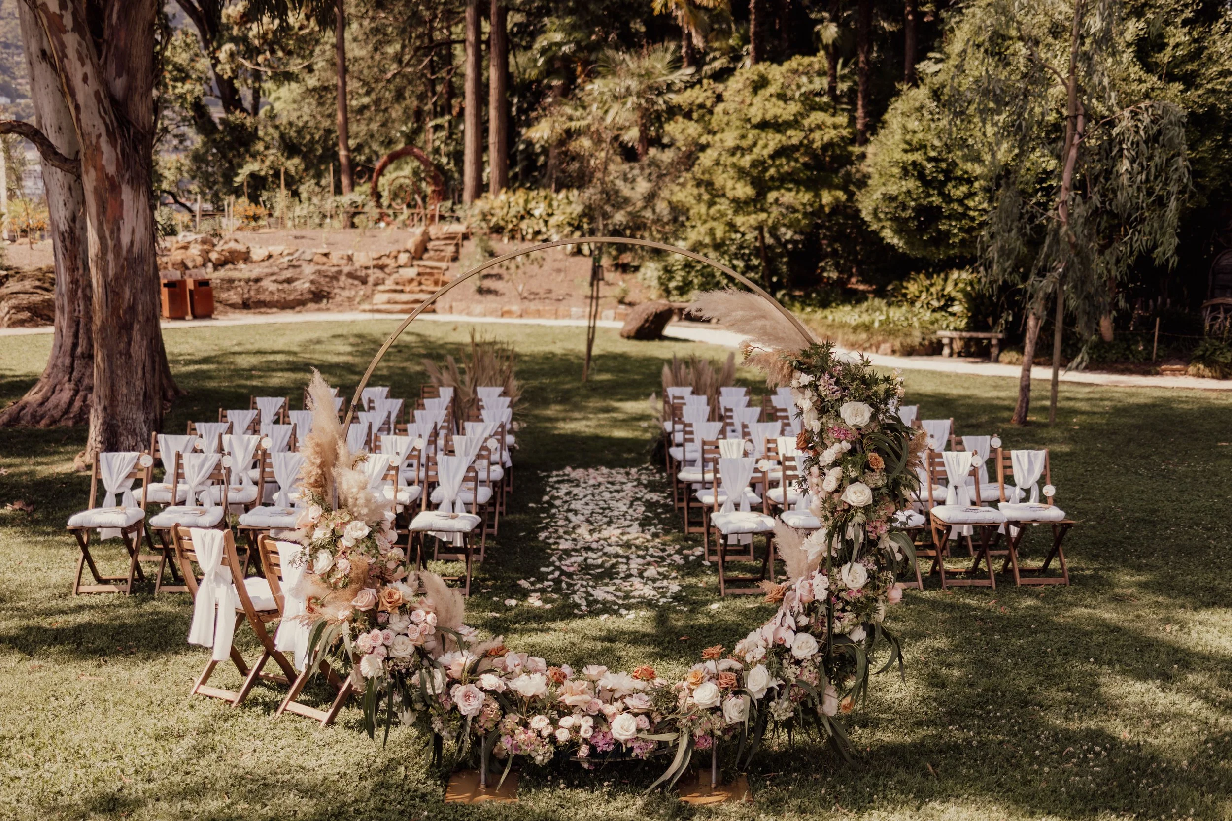 Outdoor wedding ceremony setup with rows of chairs, white floral arrangements, and a circular floral arch.