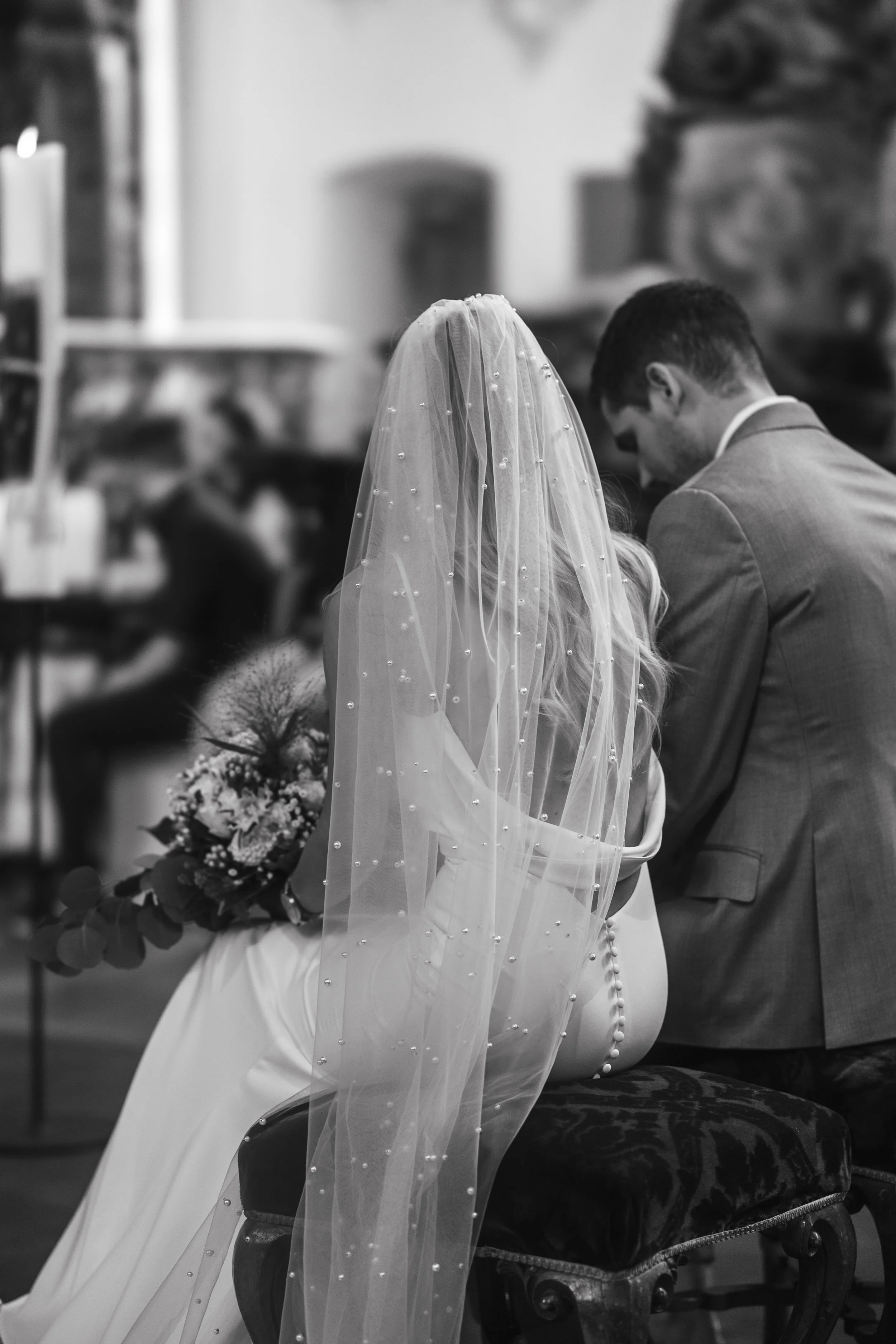 Black and white photo of a bride and groom seated during a wedding ceremony, with the bride wearing a veil and holding a bouquet.