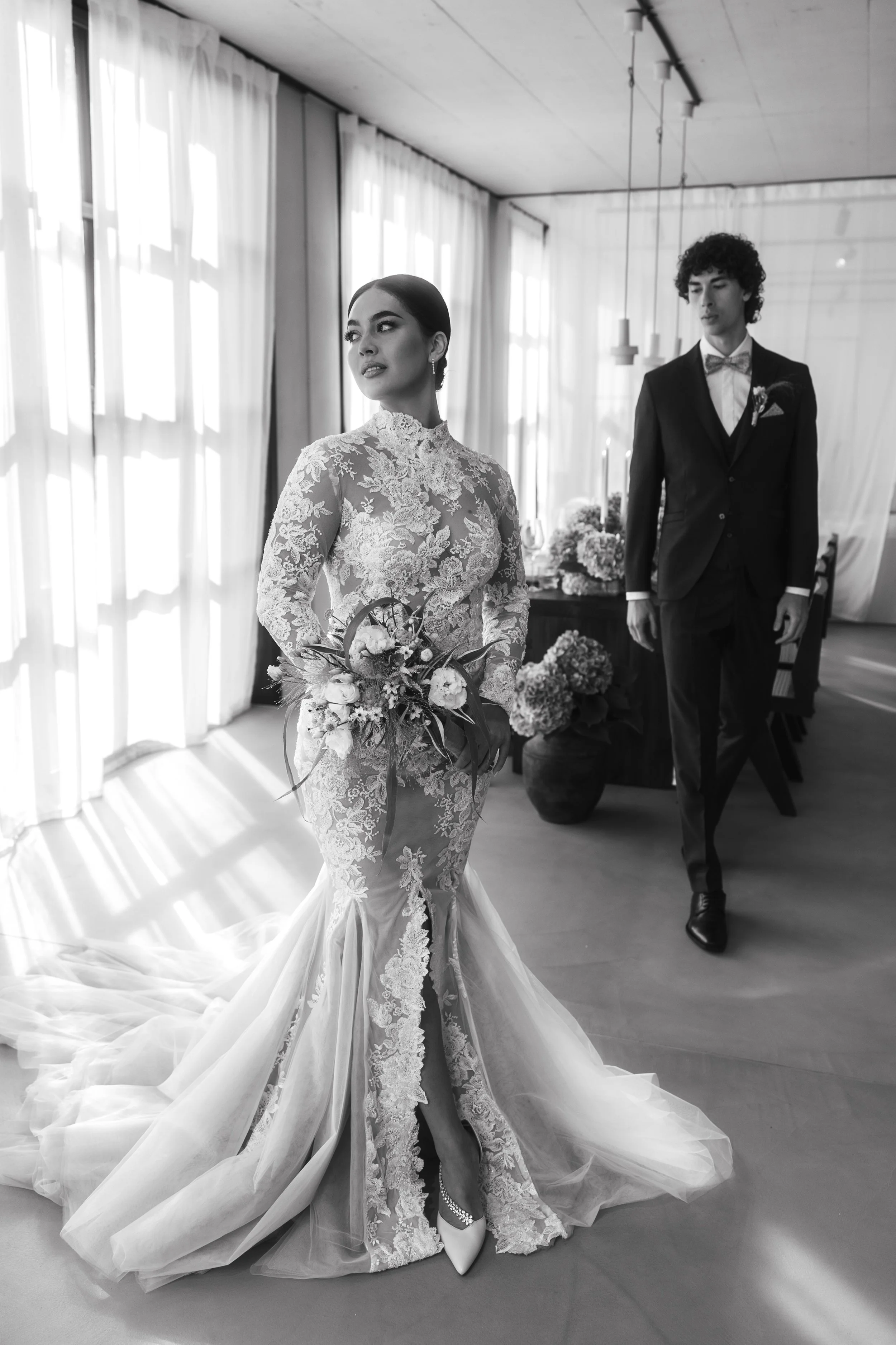 Bride in lace wedding gown holding bouquet, groom in suit behind, elegant indoor setting, black and white photo.