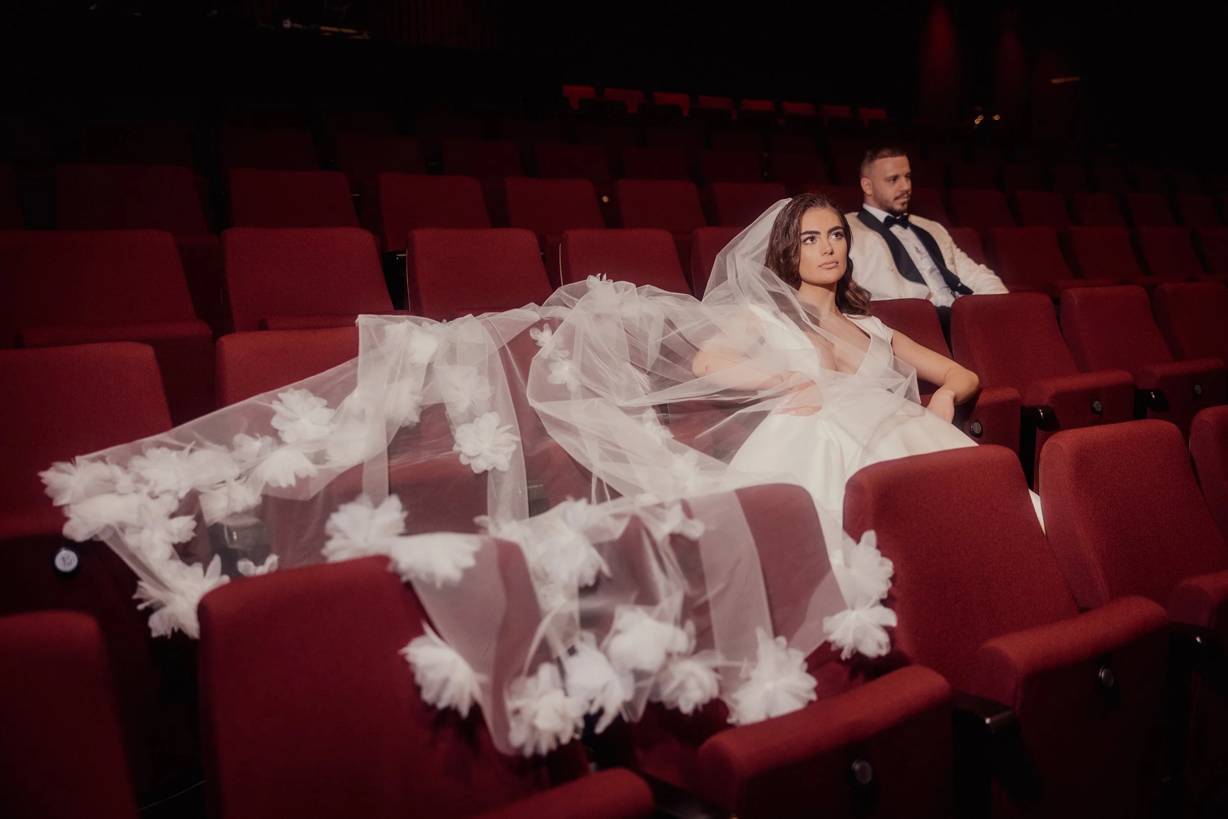 Bride in wedding dress with long veil and groom in tuxedo seated in a theater, surrounded by empty red seats.