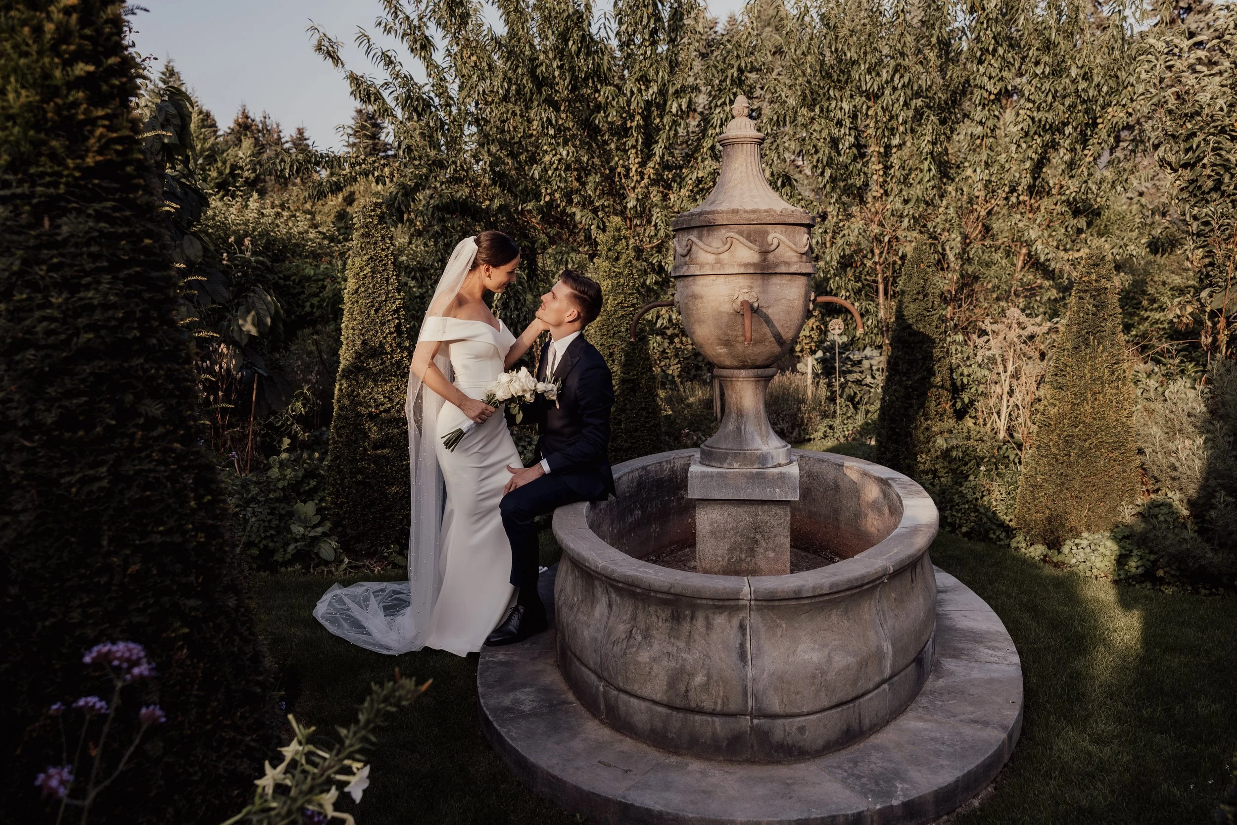 A bride and groom by a stone fountain in a garden, surrounded by greenery. The bride wears a white gown, holding a bouquet, while the groom is in a dark suit.