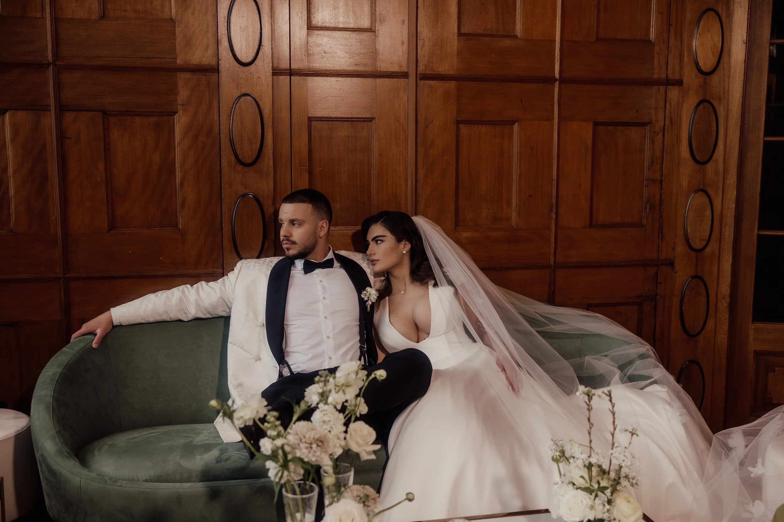 Bride and groom sitting on a green sofa, bride in a white dress with veil, groom in a white tuxedo jacket with bow tie, in front of a wooden paneled wall, floral arrangement in foreground.
