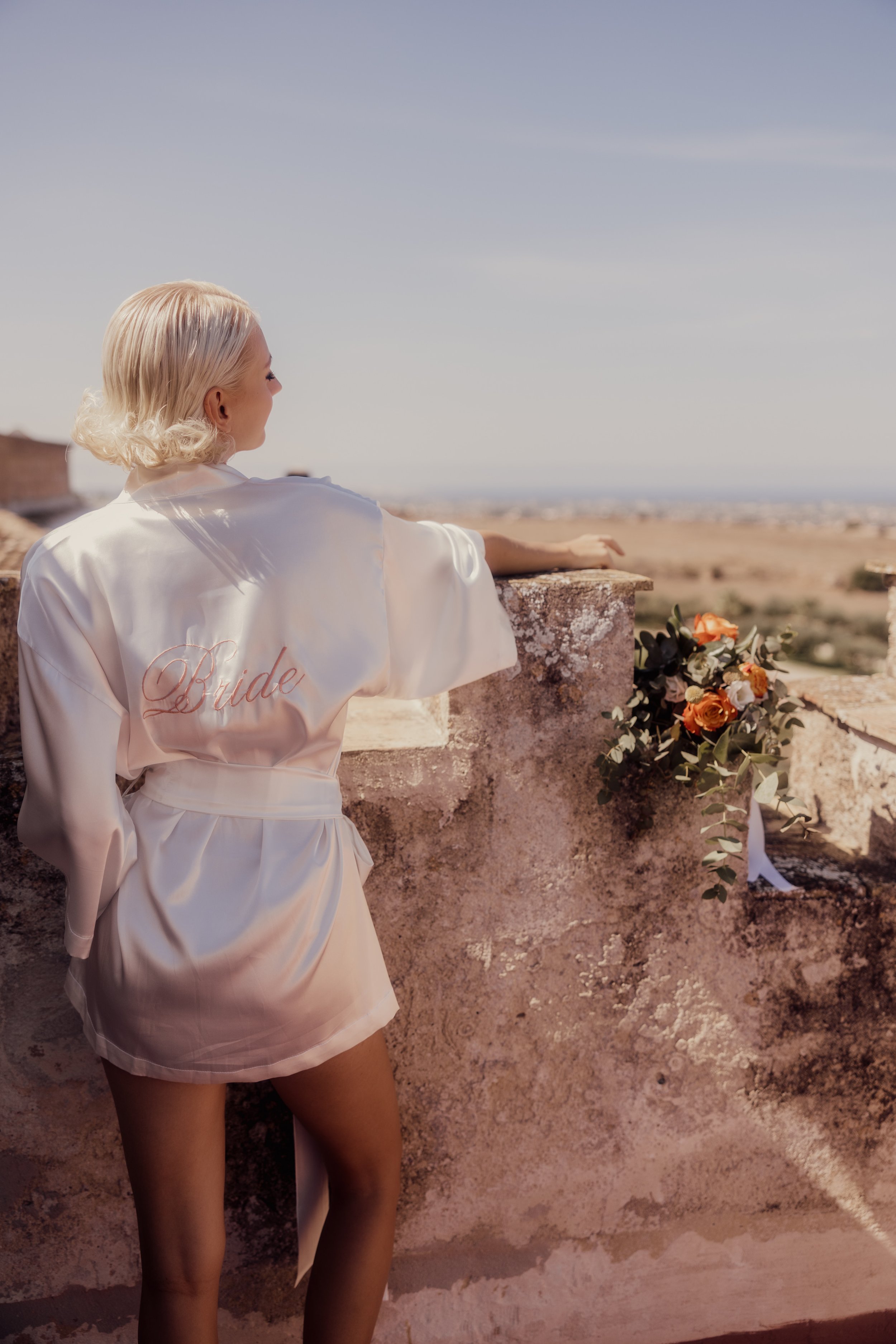 A woman in a silk robe with the word "Bride" stands at a stone wall, looking at a scenic landscape. A bouquet of orange flowers rests on the wall.