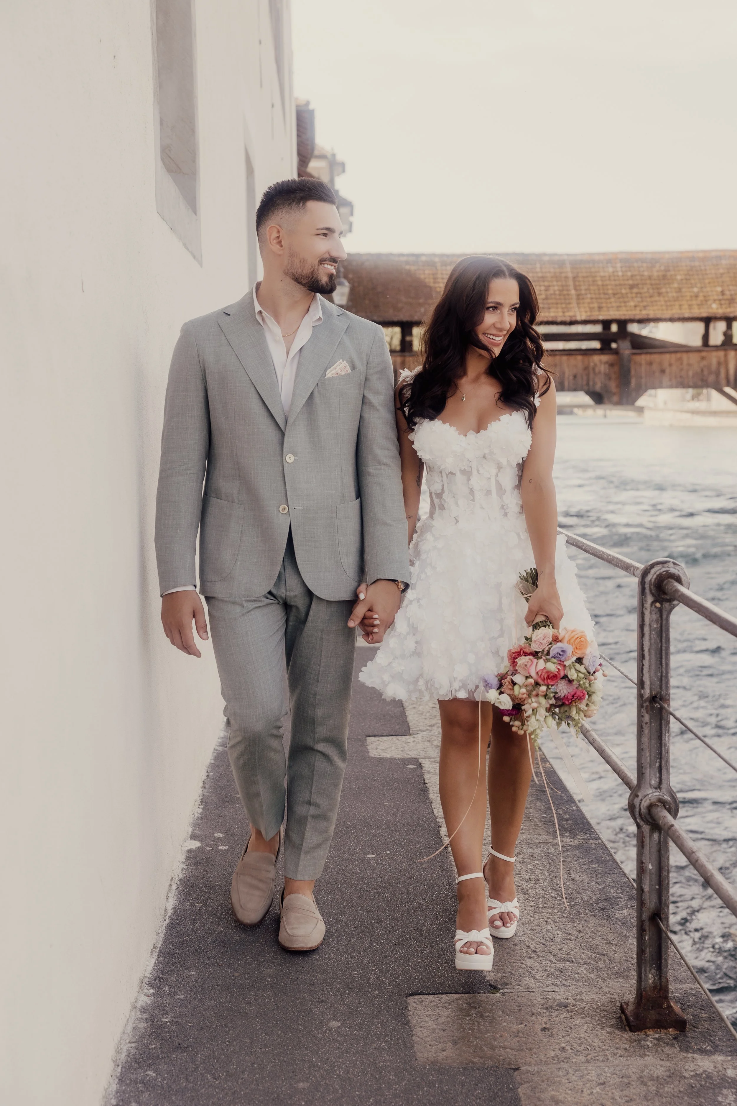 A couple walking hand in hand by a waterfront. The man is wearing a light gray suit and loafers, while the woman is in a white floral dress holding a bouquet of mixed flowers. The background features a wooden bridge over water.