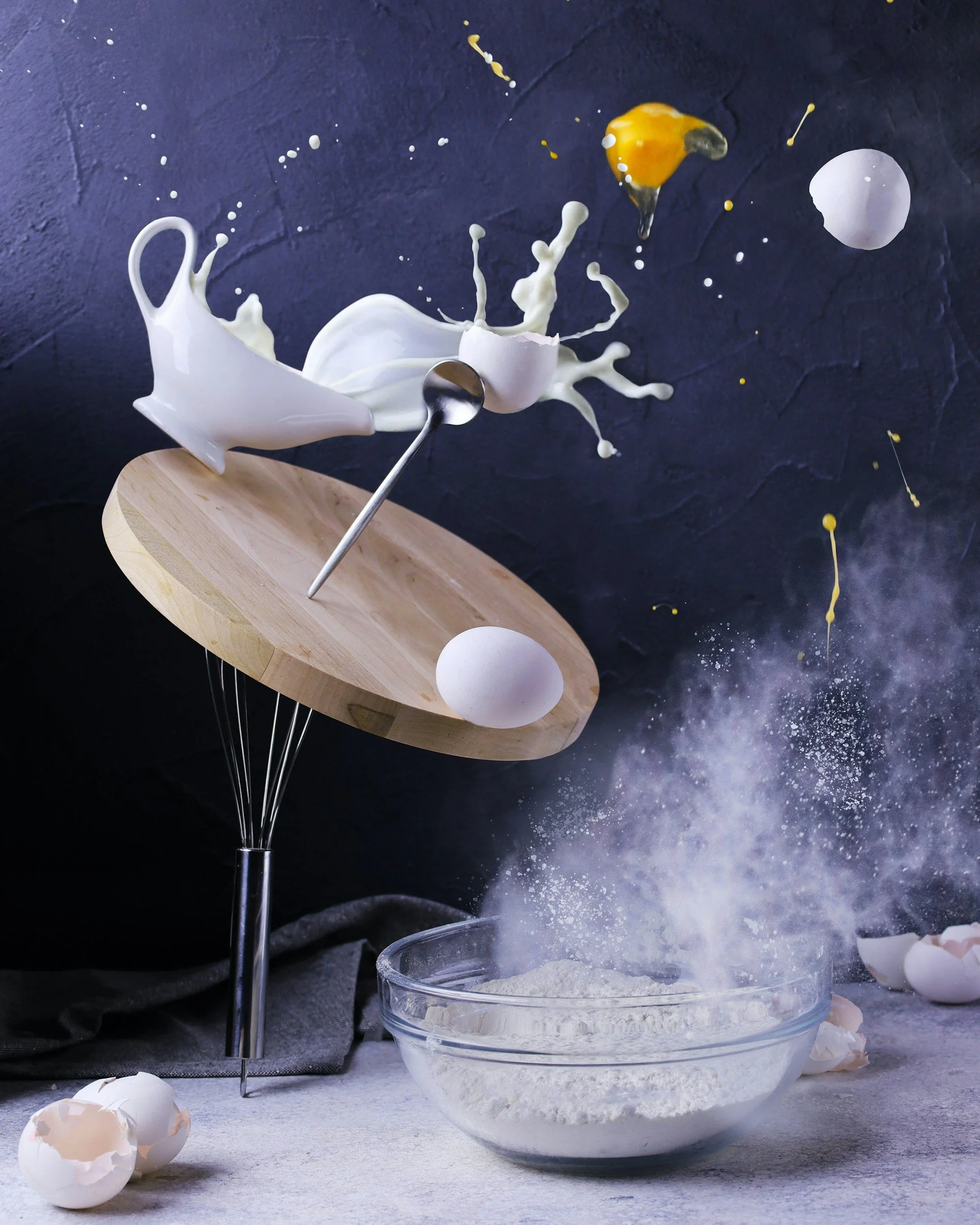 Egg and flour being thrown in the air with splashes of milk, with broken eggshells on a surface and a bowl of flour below against a dark background.