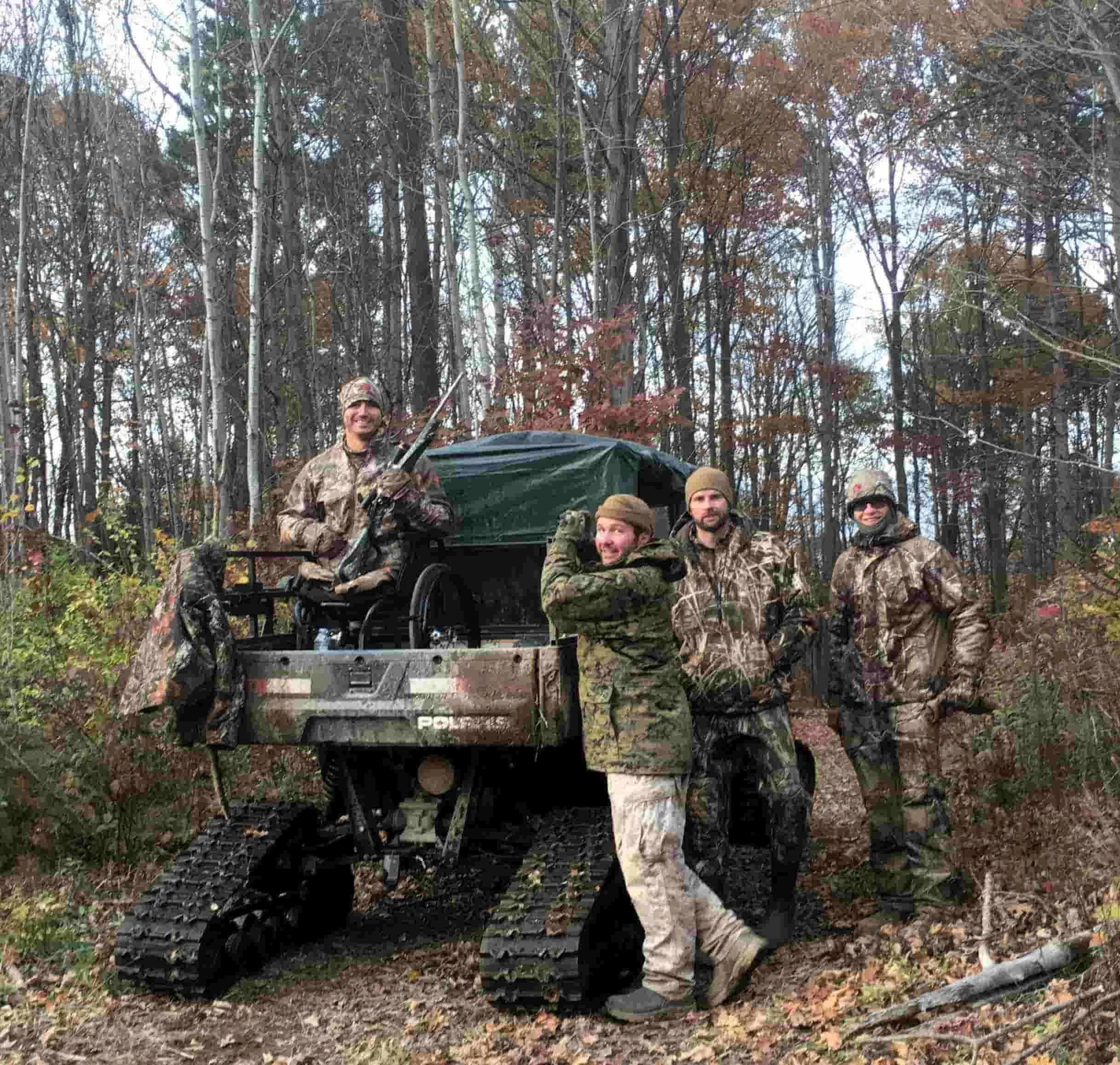 Veterans in camouflage gear standing beside an all-terrain tracked vehicle during an NPLB Outdoors adaptive hunt in the woods.