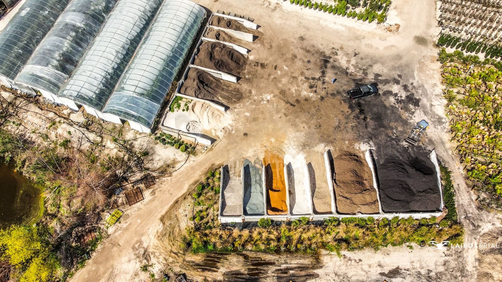 Aerial view of an industrial area with multiple greenhouses covered in transparent roofs on the left. Adjacent to the greenhouses are several large piles of different materials, possibly soil, sand, and gravel, separated into designated sections. An empty, dusty space surrounds the storage area, with a vehicle and some equipment visible. There are also rows of plants and trees. The scene is natural and industrial, indicating a plant nursery or landscaping supply facility.