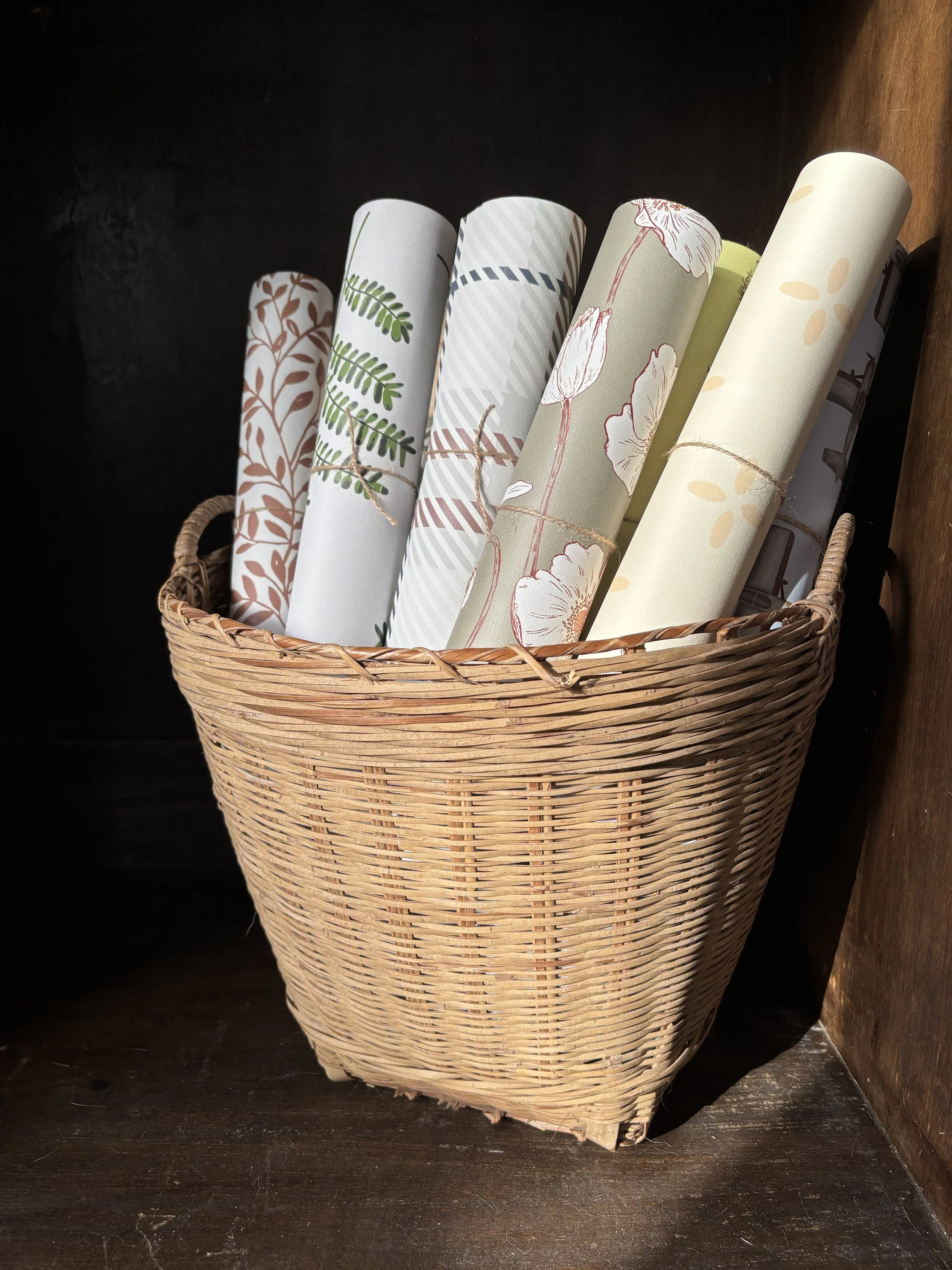 A basket filled with rolled wallpaper with various floral and leaf patterns, placed on a dark wooden surface with a dark background.