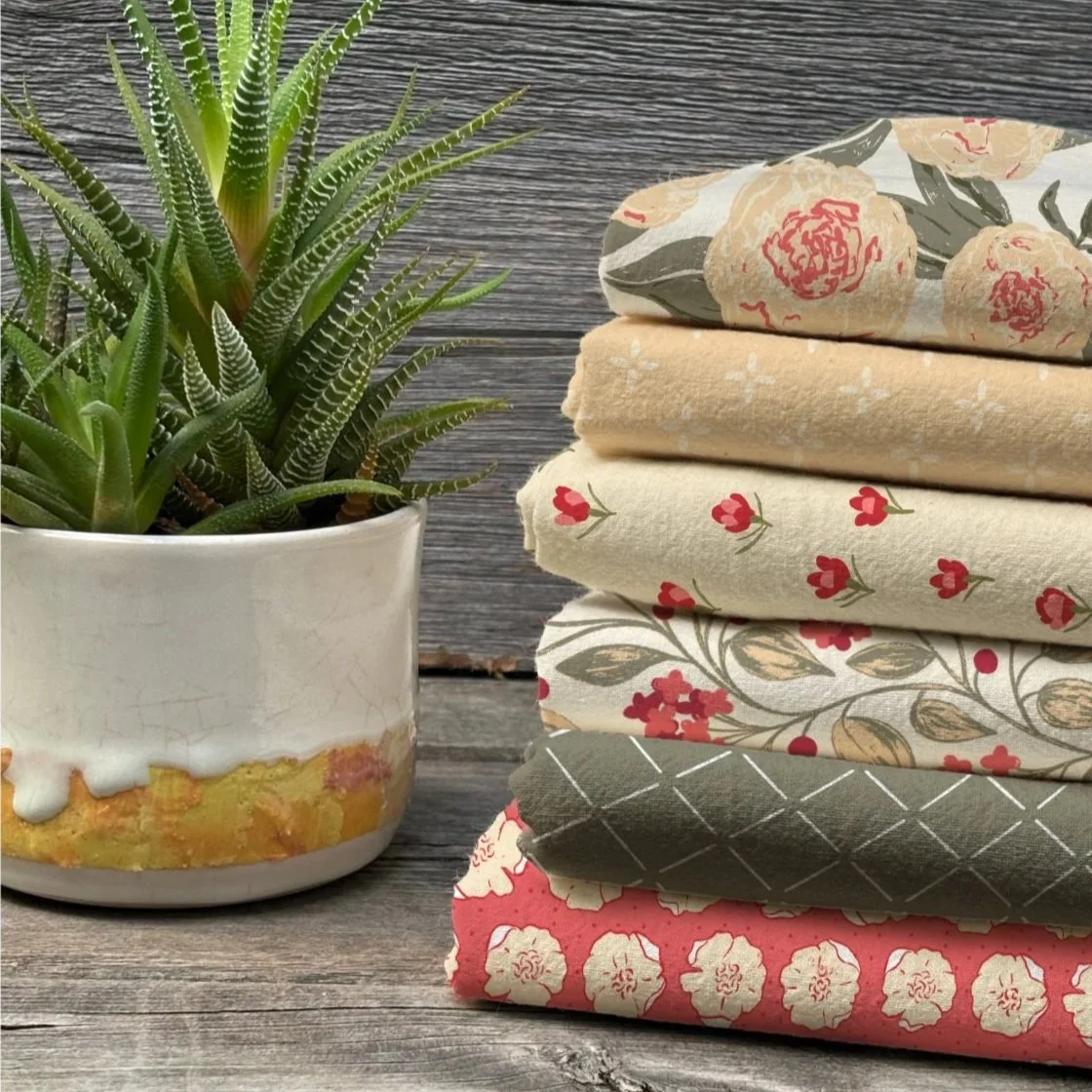A potted succulent plant with spiky green leaves next to a stack of six folded fabrics with floral and geometric patterns, on a rustic wooden surface with a wooden background.