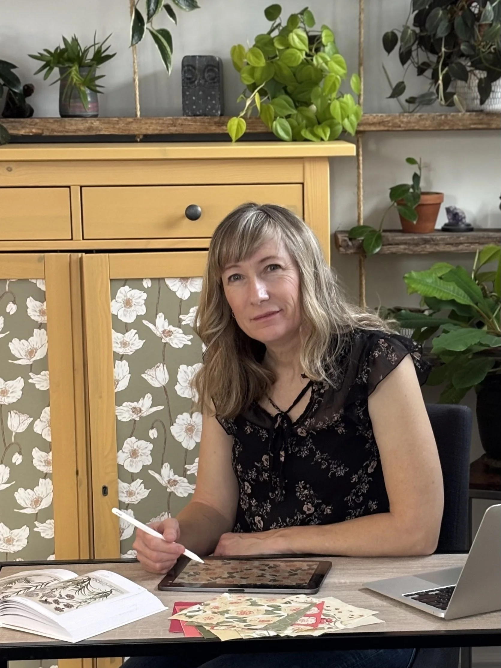 A female artist creating a repeating pattern at a desk with a laptop, tablet, open book, and decorative paper, surrounded by green plants on shelves behind her.