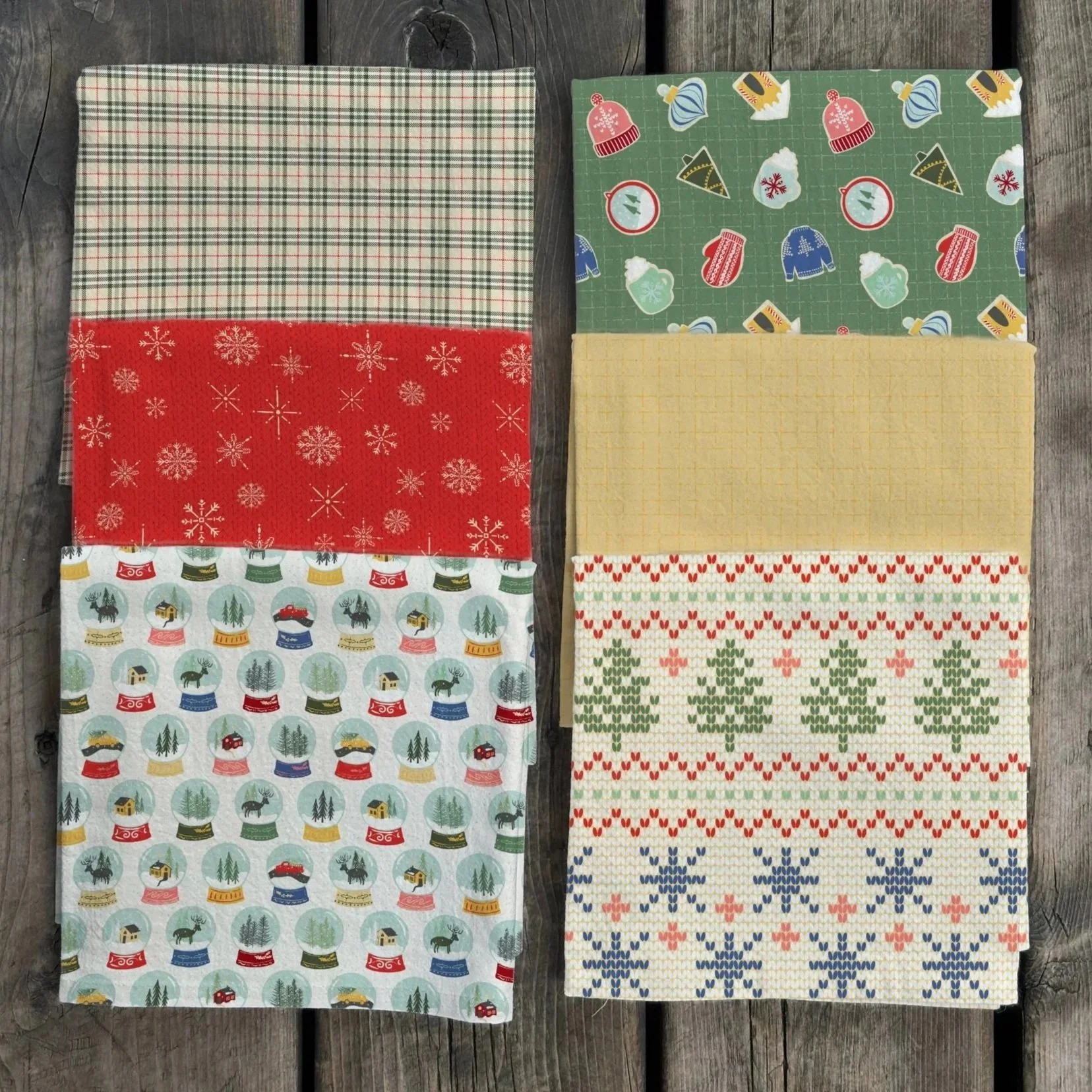 A collection of Christmas-themed fabrics with various festive patterns, including snowflakes, Christmas trees, snow globes, and holiday icons, arranged on barnboard surface.