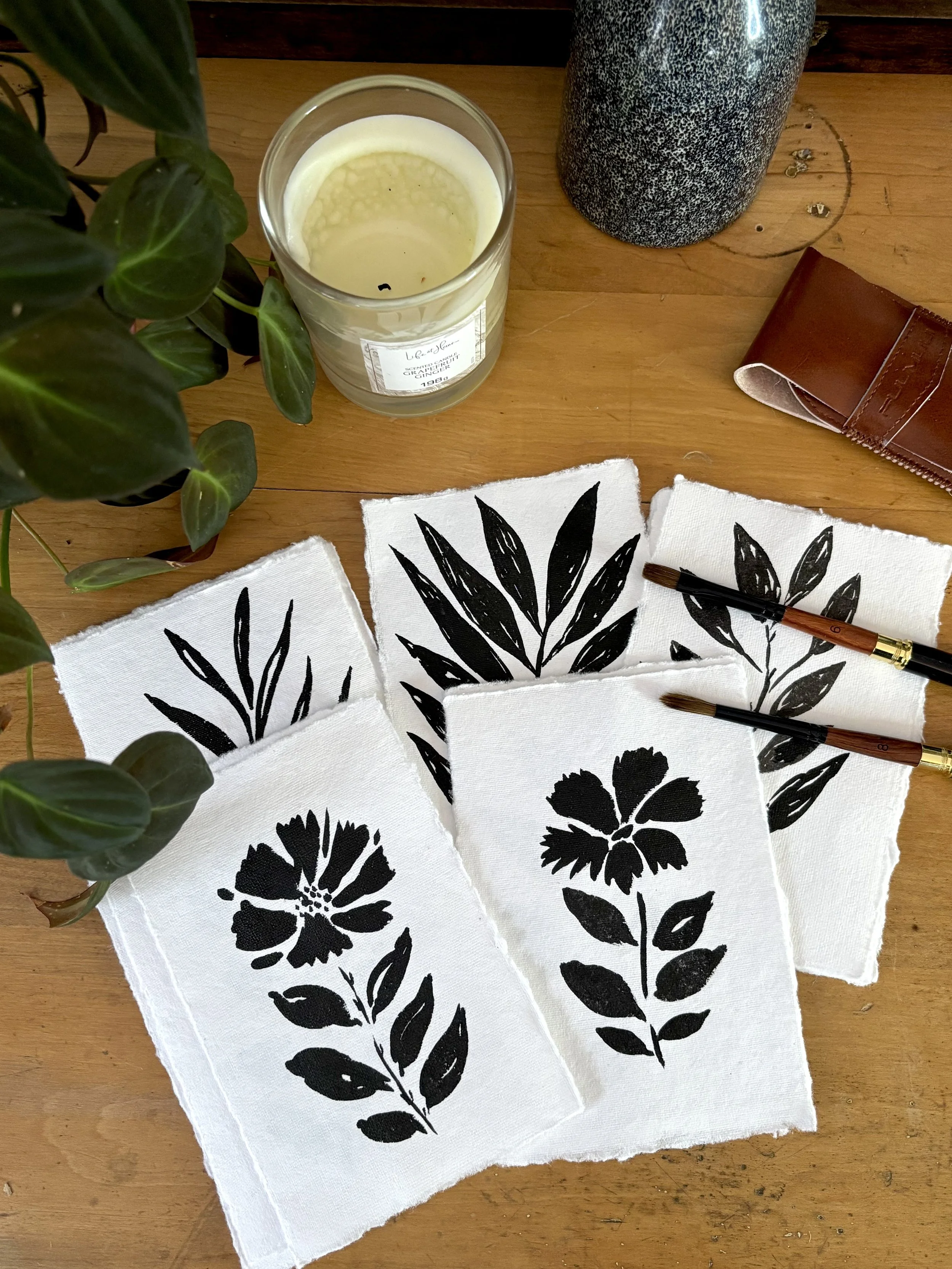 Black ink paintings of botanicals, flowers and leaves, on a wooden table. Two paintbrushes rest on two of the paintings. To the top is a candle, a plant, and a gray ceramic vase.  