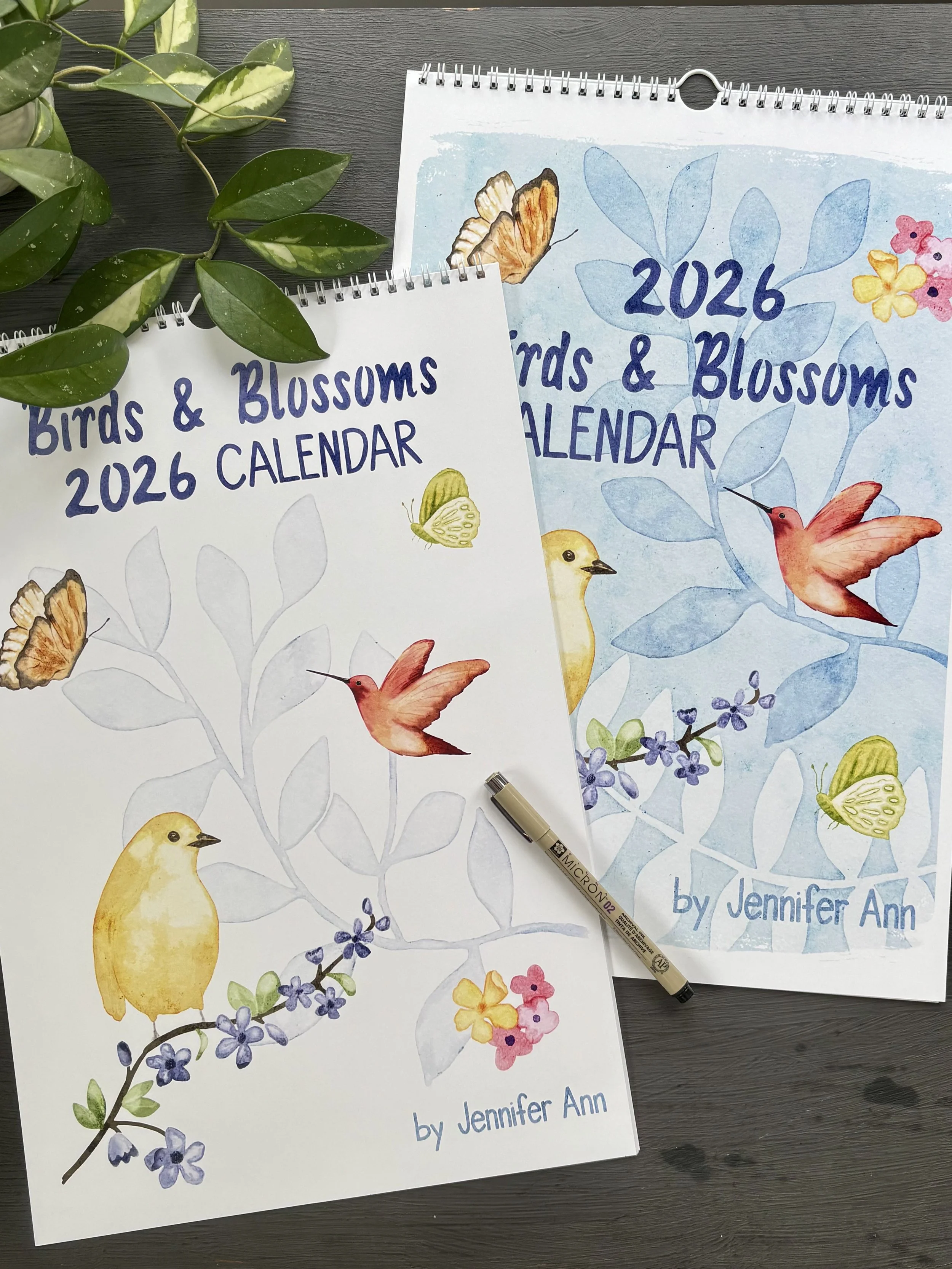 Two 2026 Birds & Blossoms calendars with watercolor illustrations of birds, butterflies, and flowers, placed on a dark wooden surface with a pen and green plants nearby.