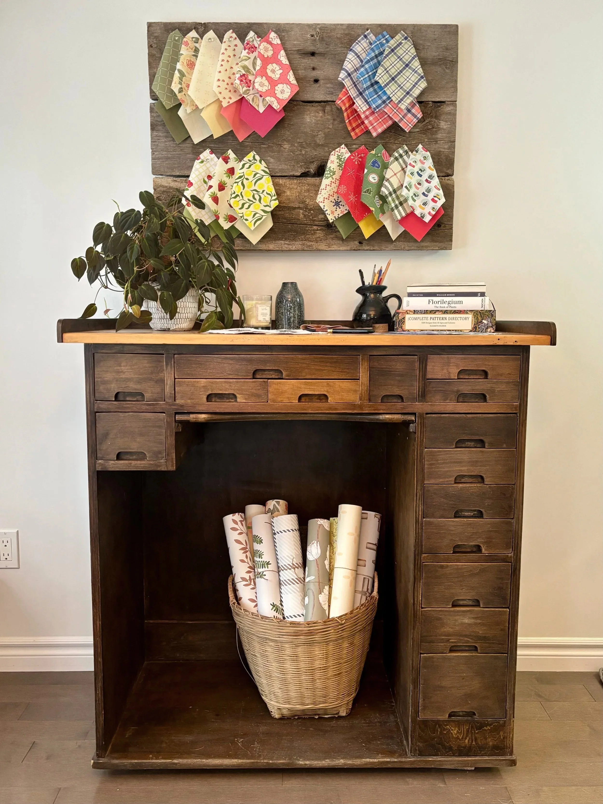 An image of a studio workspace. A vintage jewelers table with multiple small drawers with a basket of patterned wallpaper, and a rustic wall display of fabric swatches above.