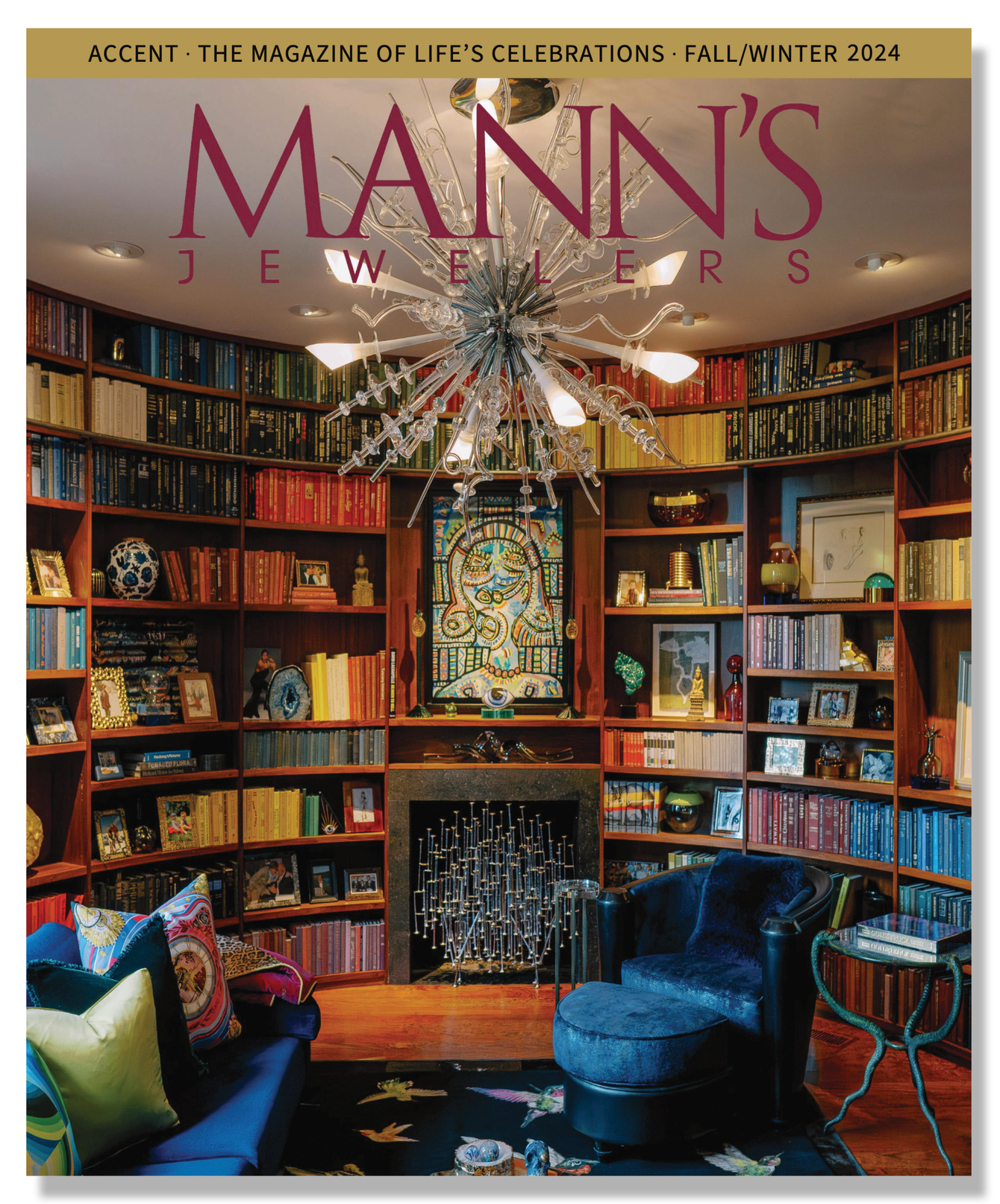 Inside a luxurious library with a built-in bookshelf filled with colorful books decorated with artwork and photographs. A modern chandelier hangs on the ceiling, and there's a fireplace with a unique sculpture on top. The room has dark plush armchairs and a sofa with decorative pillows, and a patterned rug on the wooden floor.