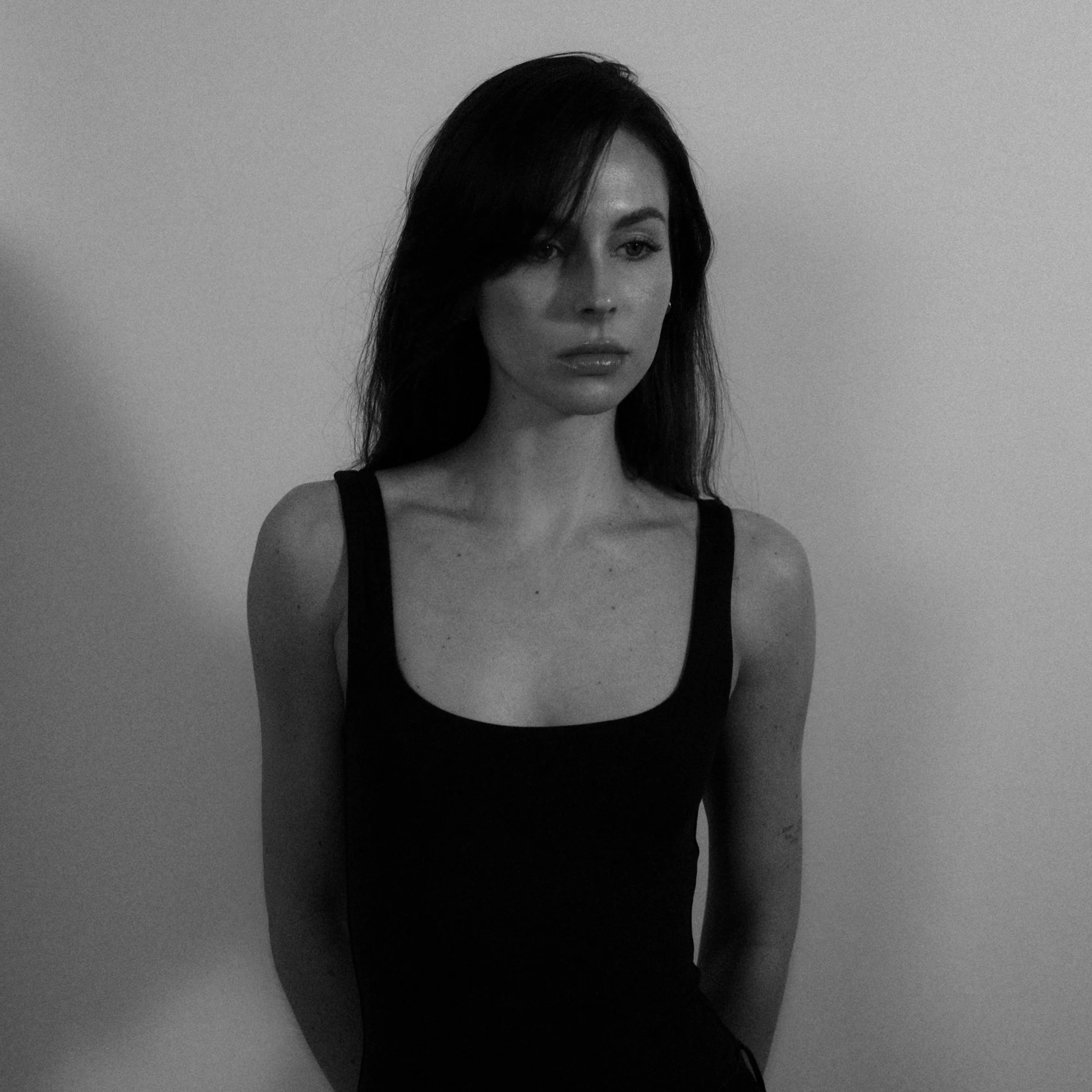 Black and white photo of a woman with long dark hair wearing a sleeveless black top, standing against a plain wall.