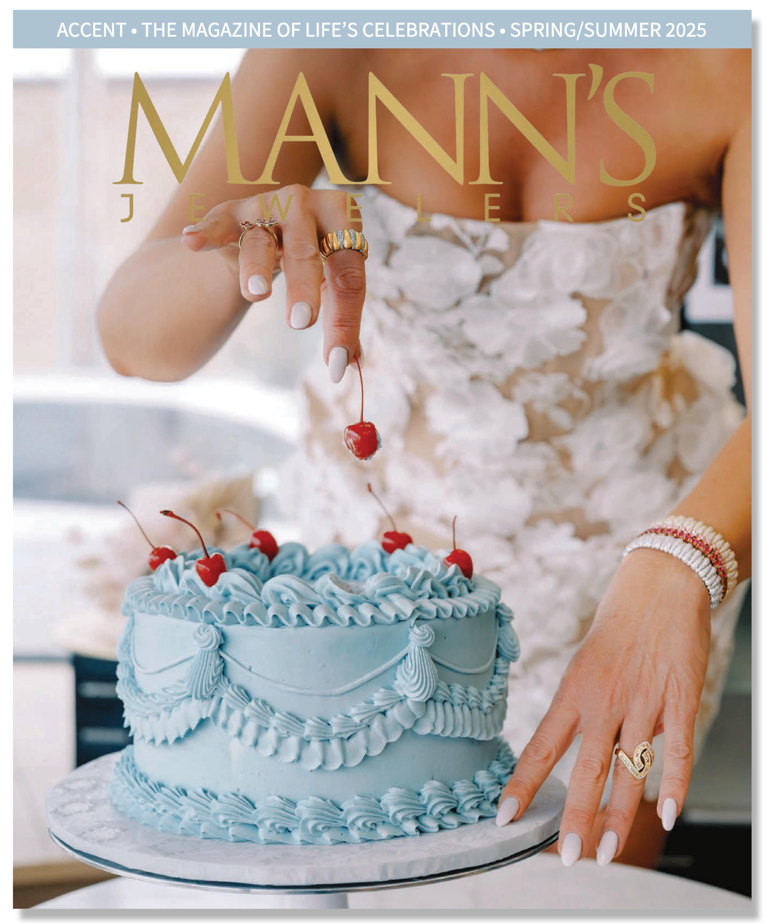 A woman in a white, textured dress is placing a cherry on a blue frosted cake with several cherries on top. She is wearing rings and bracelets on her right hand and has neatly manicured white nails.