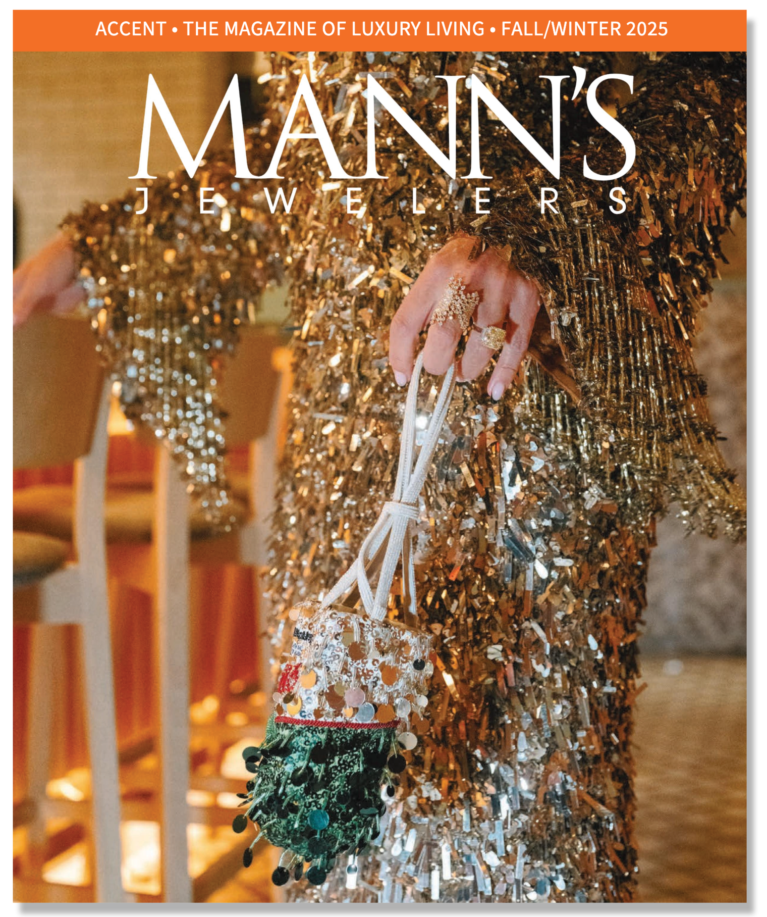 A person wearing a sequined, gold-colored outfit holding a decorated handbag, with a background of chairs and a table, featured on the cover of Mann's Jewelers Fall/Winter 2025 magazine.