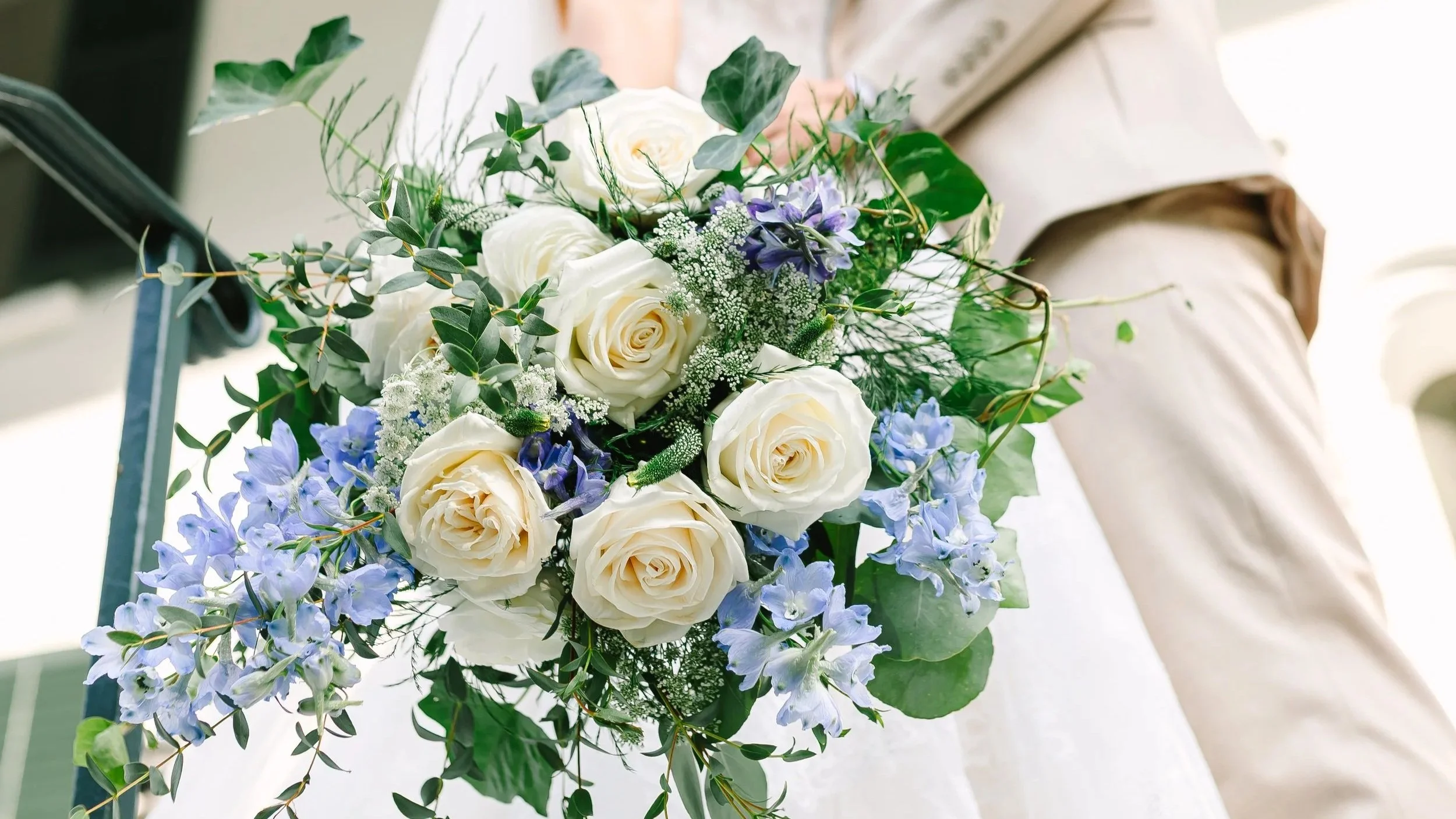 A bridal bouquet with white roses, purple flowers, and green foliage held by a person in a beige or light-colored dress.