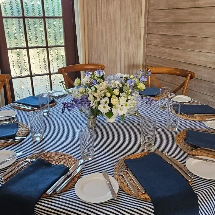 Round dining table set for a meal with navy blue napkins, clear glasses, white plates, and a floral centerpiece with white and purple flowers, in a room with wooden walls and a window.