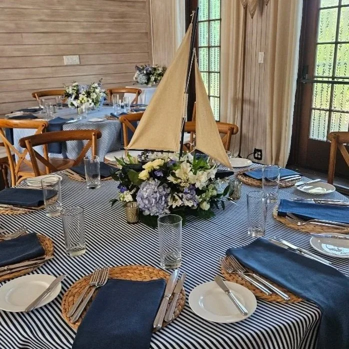 Round dining table with a navy and white striped tablecloth, set with white plates, silverware, napkins, and empty glasses. The centerpiece is a floral arrangement with white and purple flowers and a small sailboat model. Wooden chairs surround the t