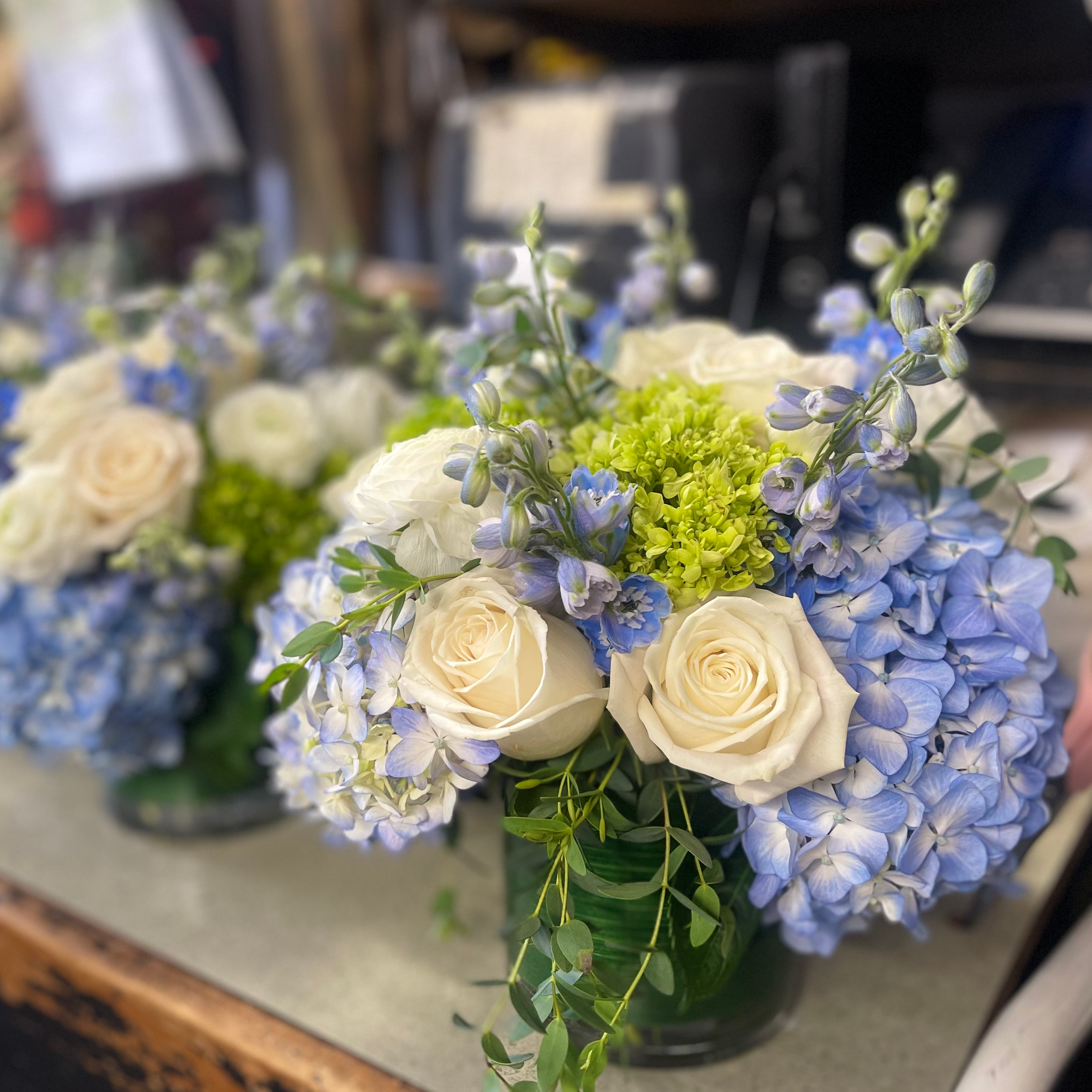A bouquet of white roses, blue hydrangeas, and green foliage in a vase, placed on a table.