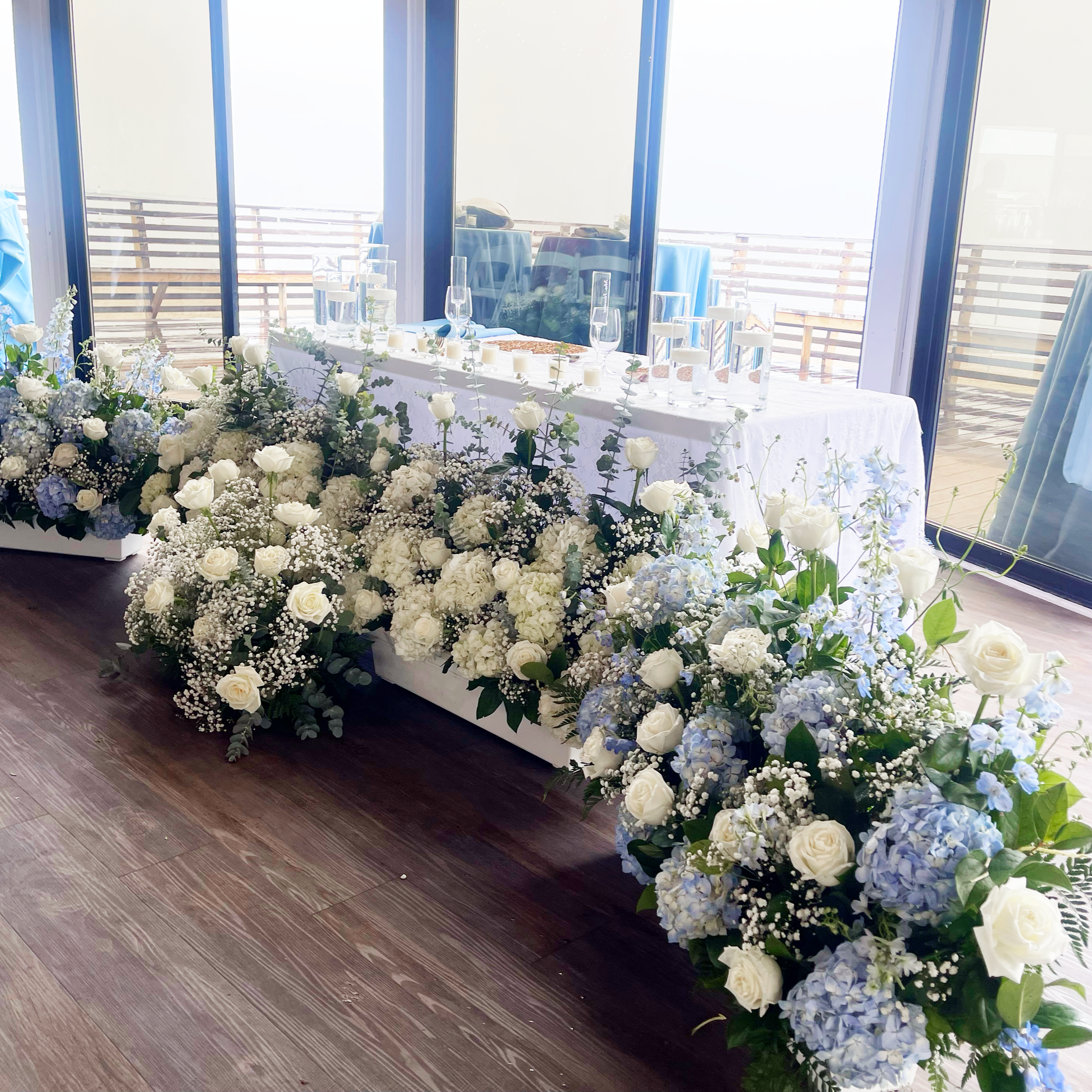 A beautifully decorated table with white flowers, candles, and glasses set up for an event, with large windows and an outdoor balcony in the background.