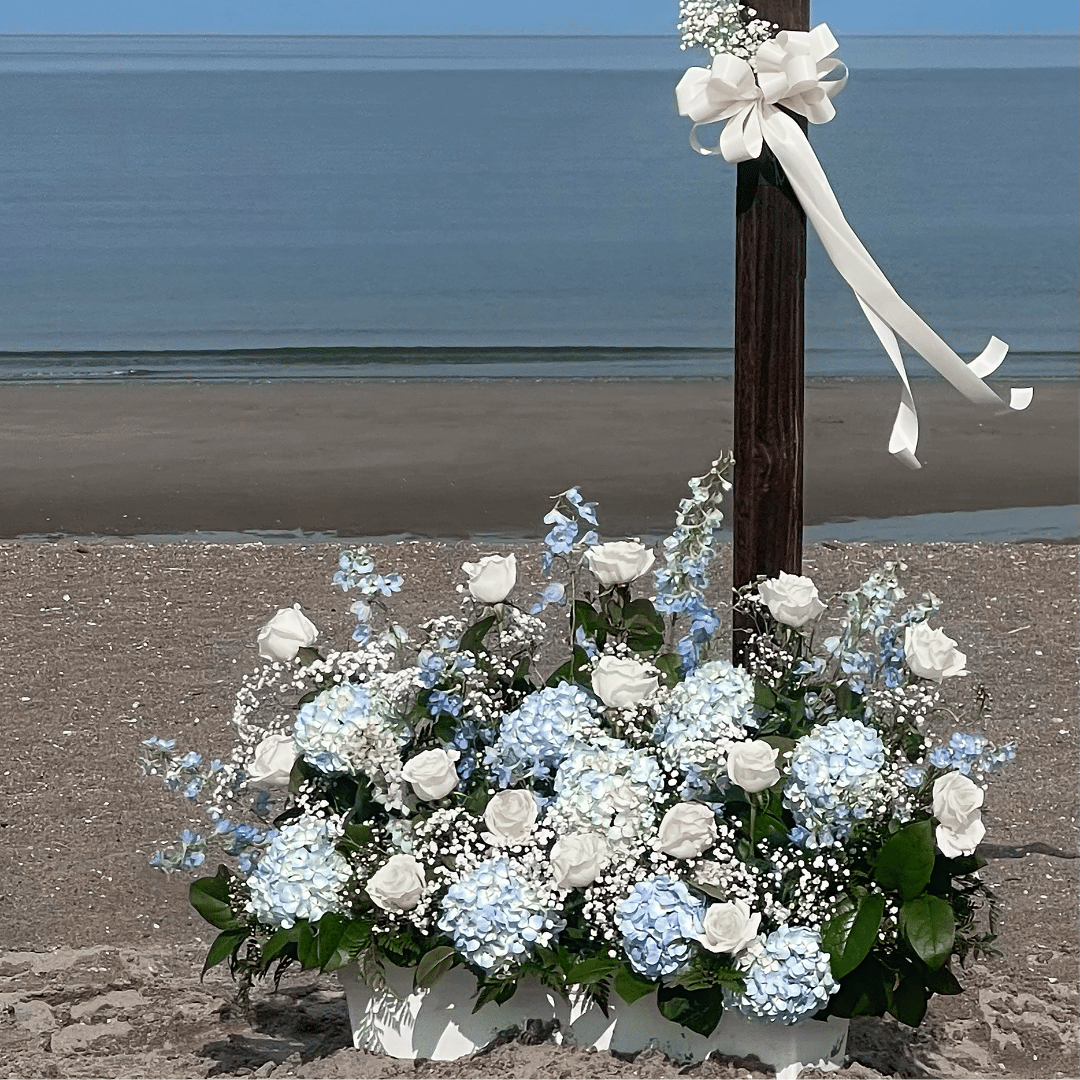 A wedding flower arrangement with white roses and hydrangeas on the beach near the ocean, decorated with a white ribbon and bow tied on a wooden post.