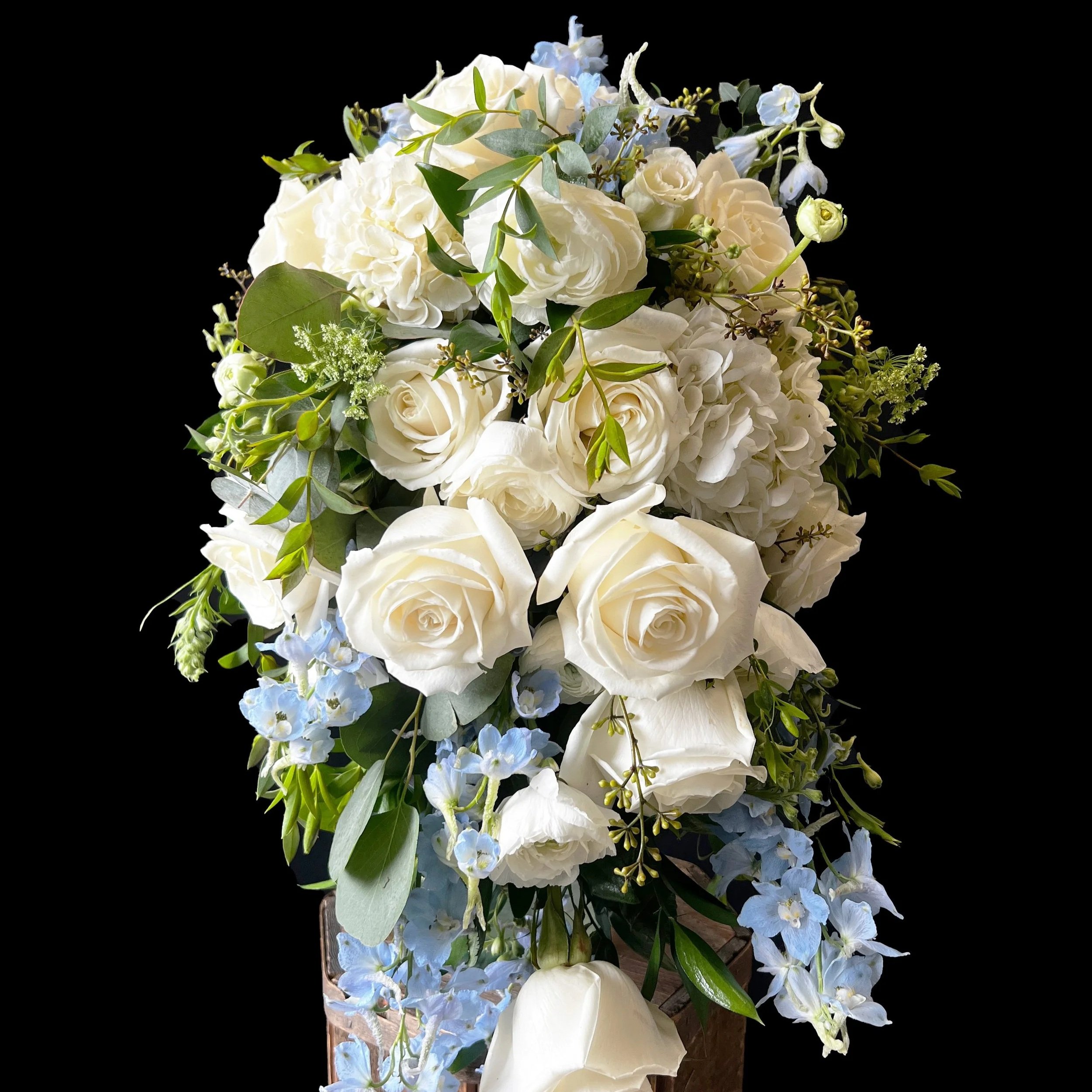 A wedding bouquet consisting of white roses, hydrangeas, blue delphiniums, and green foliage, arranged in a tall, cascading style.