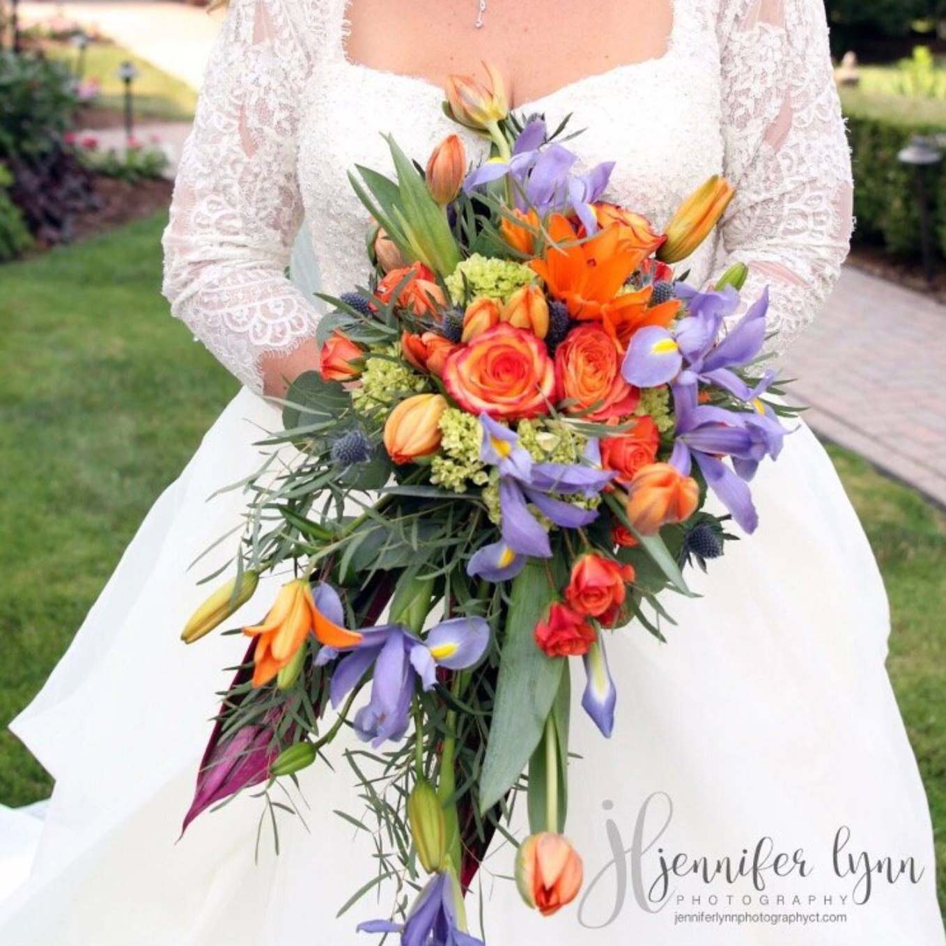 A bride in a white wedding dress holding a colorful bouquet of orange, purple, and green flowers outdoors on a garden pathway.