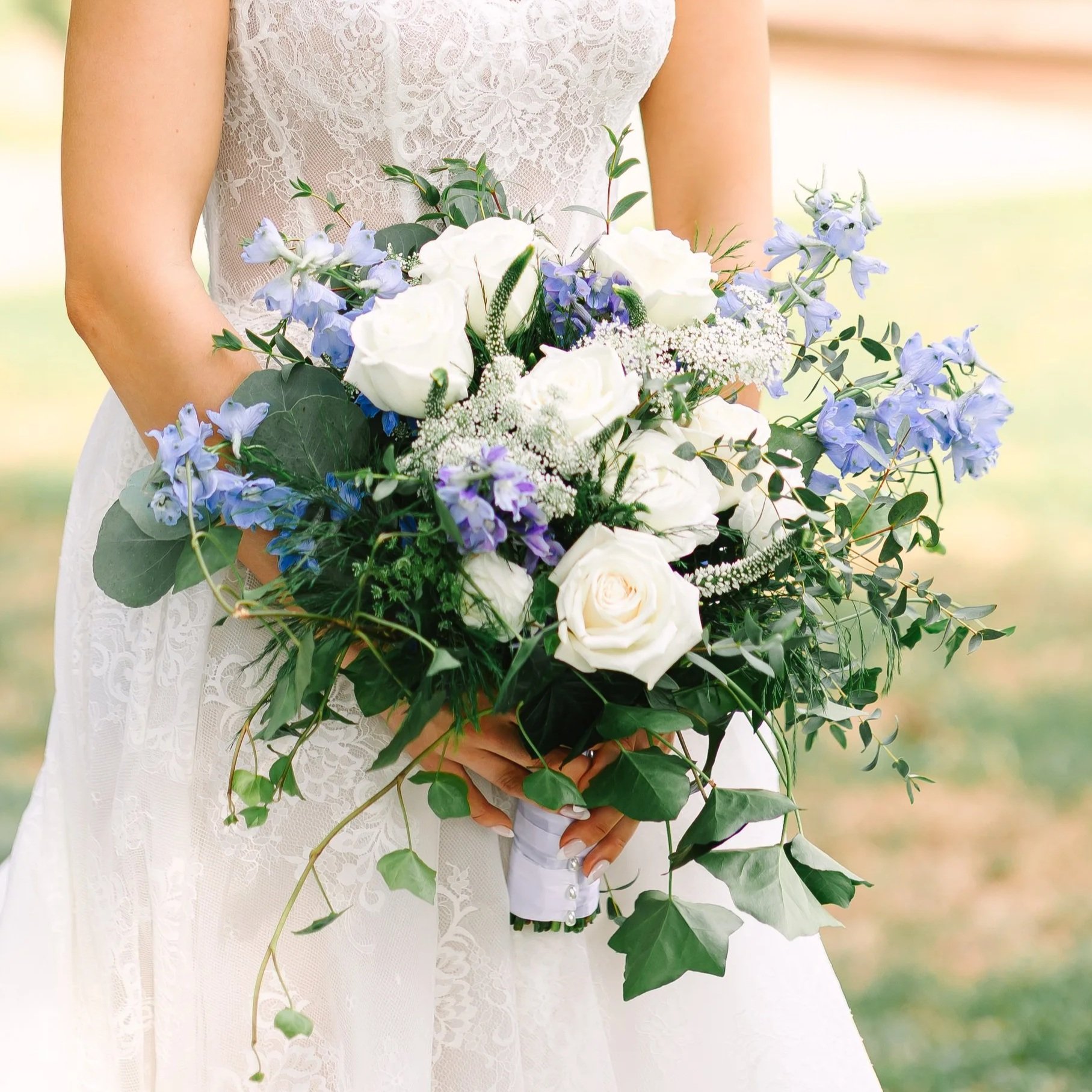 A bride in a white wedding dress holding a bouquet of white roses and blue flowers, outdoors on a sunny day.