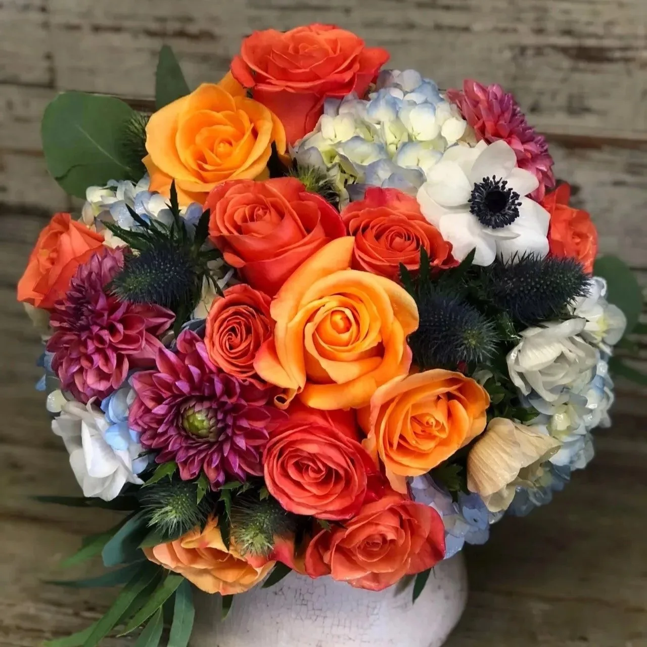 A colorful bouquet of orange, red, yellow, pink, white, and purple flowers in a white vase with a wooden background.