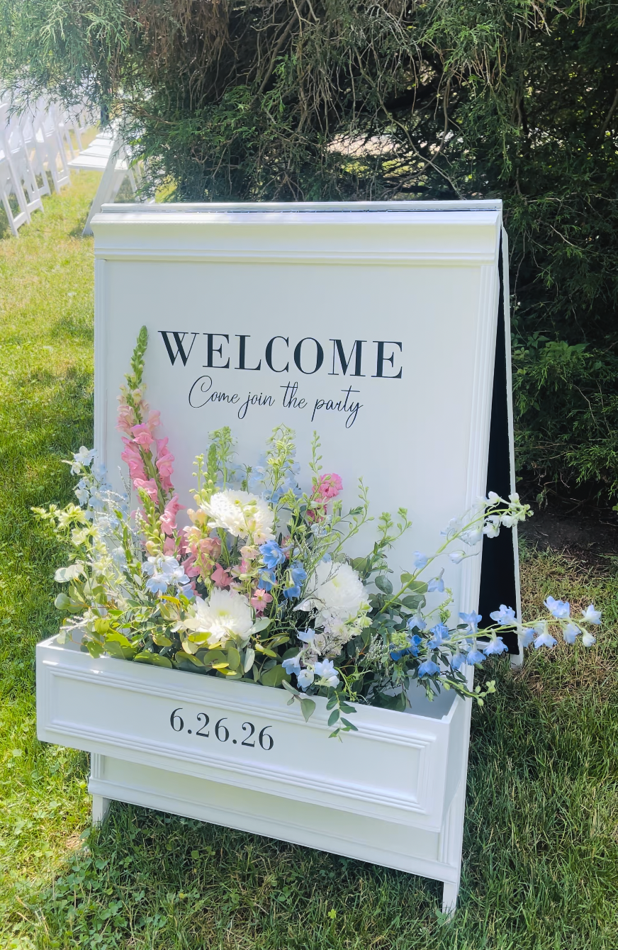 White decorated welcome sign with a flower box attached with pink, white, and blue flowers in the box. 