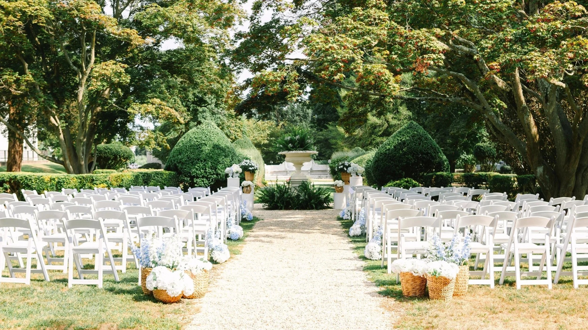 Outdoor wedding ceremony setup with white chairs lined in rows, floral arrangements, and a garden with trees and greenery in the background.
