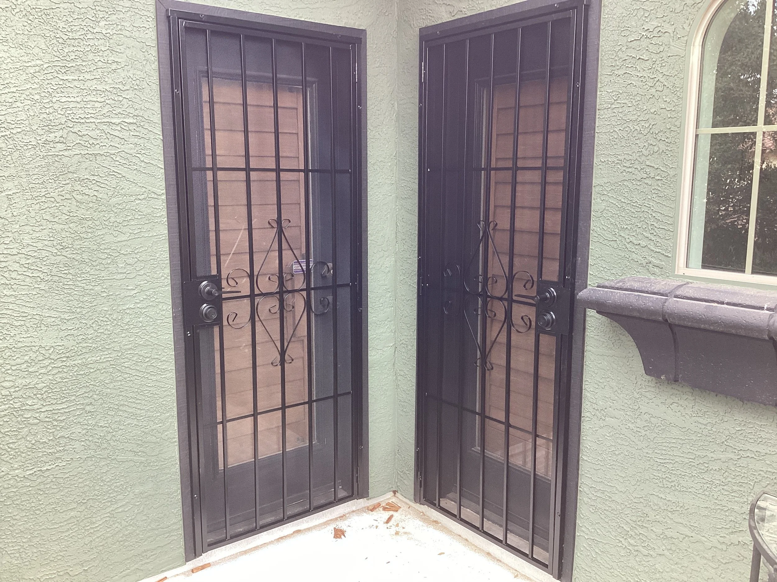 Two black screen door security doors with decorative ironwork in front of glass doors, next to a green textured wall and a window with a white frame and a black window sill.