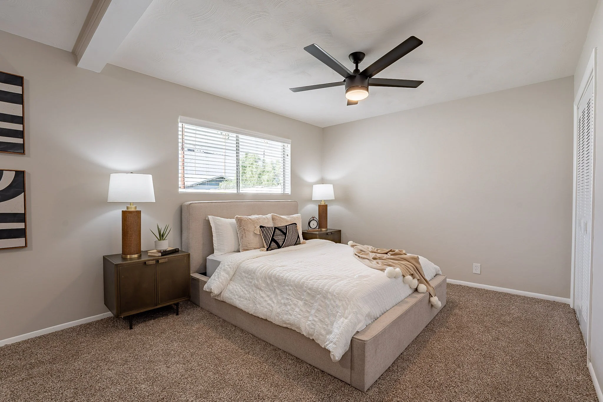 A modern bedroom featuring a beige upholstered bed with white bedding and decorative pillows, two matching nightstands with table lamps, a window with blinds, a ceiling fan, and wall art.