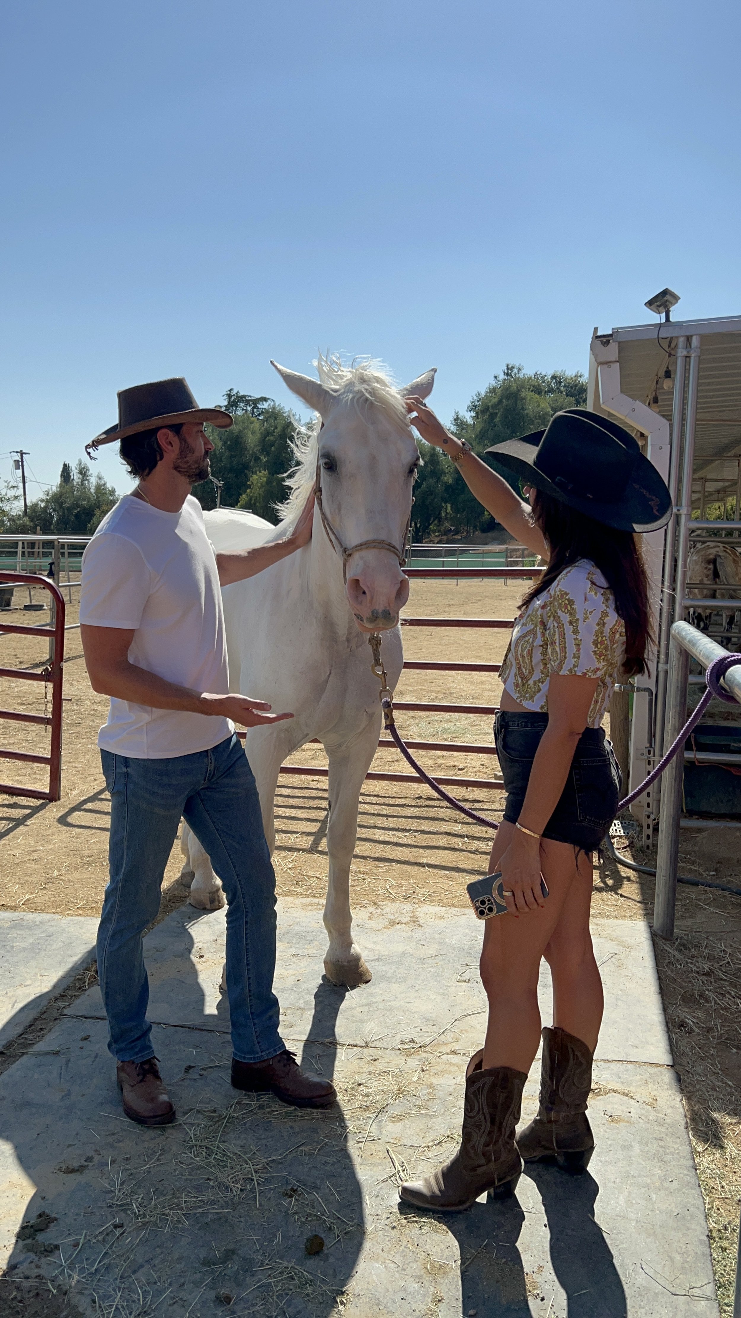 A man and woman wearing cowboy hats interacting with a white horse in a sunny outdoor setting at Ranch Reset