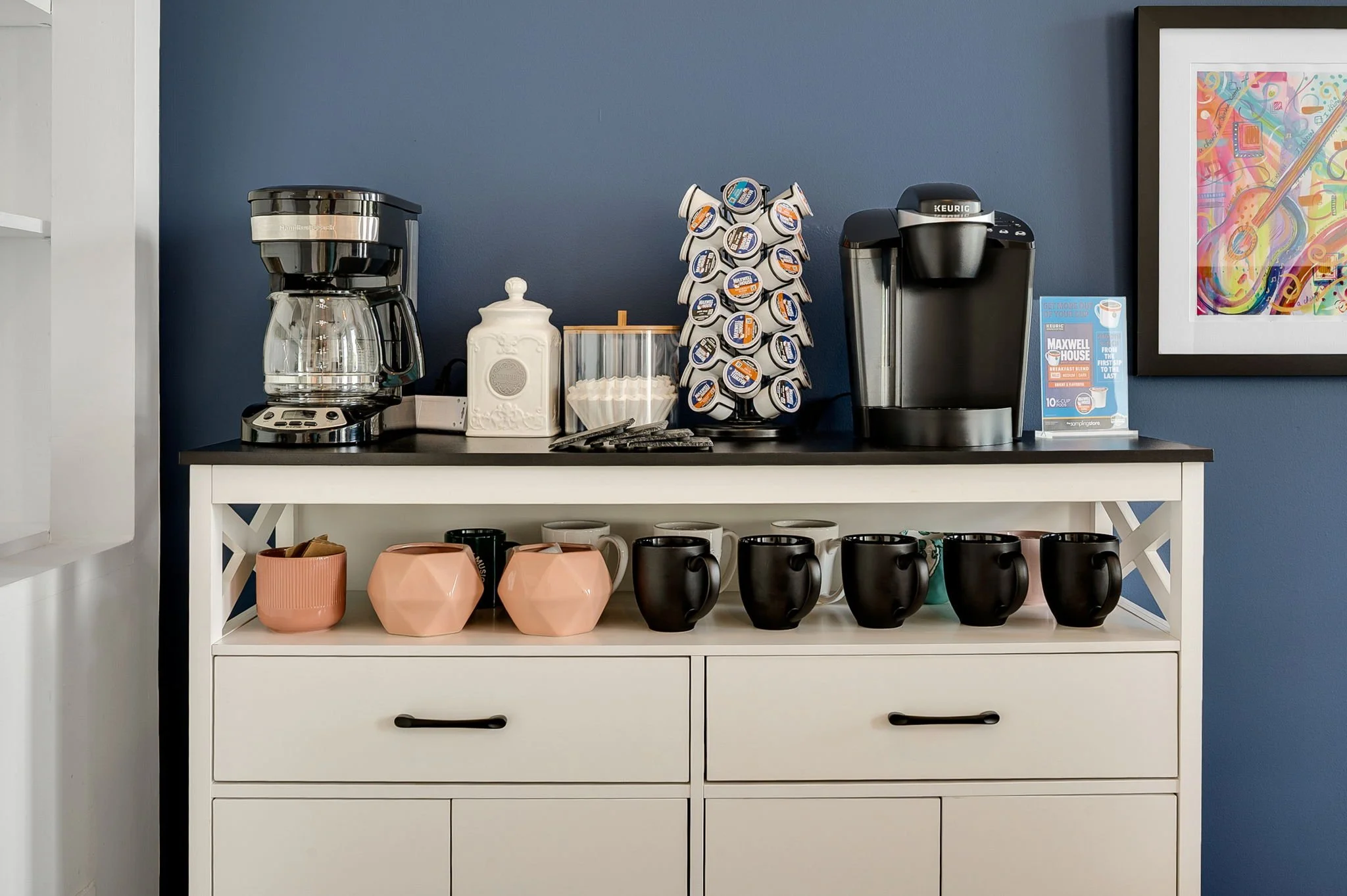 A coffee station with a coffee maker, a Keurig machine, a coffee pod carousel, cups, and decorative containers on a white cabinet with drawers, against a blue wall with colorful framed art.