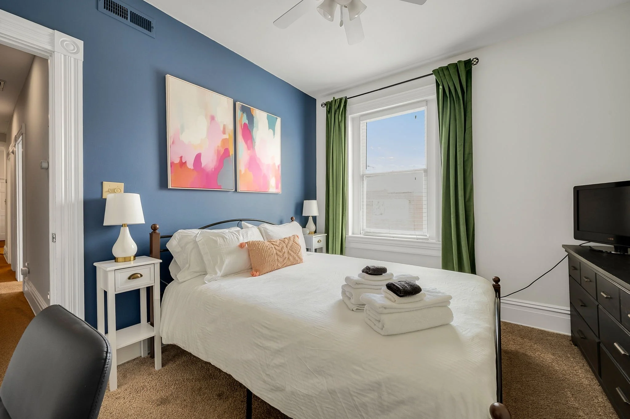 A bedroom with white walls and a blue accent wall, featuring a bed with white bedding and decorative pillows. There are two white nightstands with lamps, a window with green curtains, and a dresser with a small TV. Two abstract paintings hang on the 