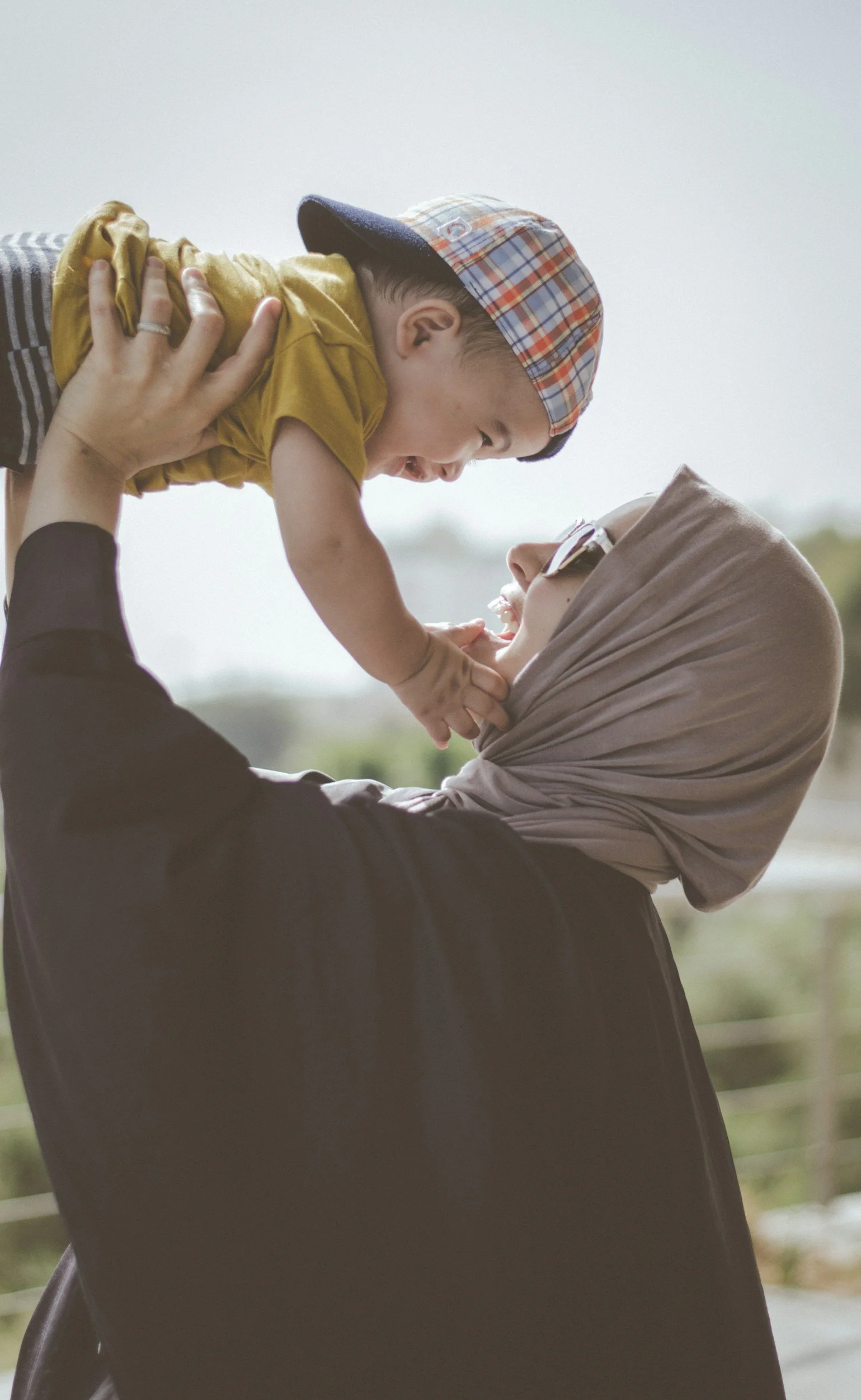 A woman in a hijab and sunglasses lifts a young boy with a plaid cap into the air, both smiling and facing each other, outdoors on a cloudy day.
