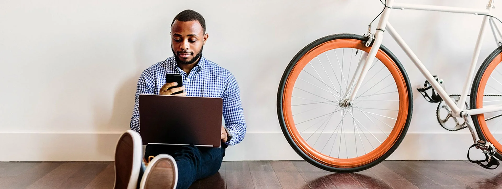 A man sitting on the floor using a laptop and looking at his phone, with a white bicycle with orange tires in the background.