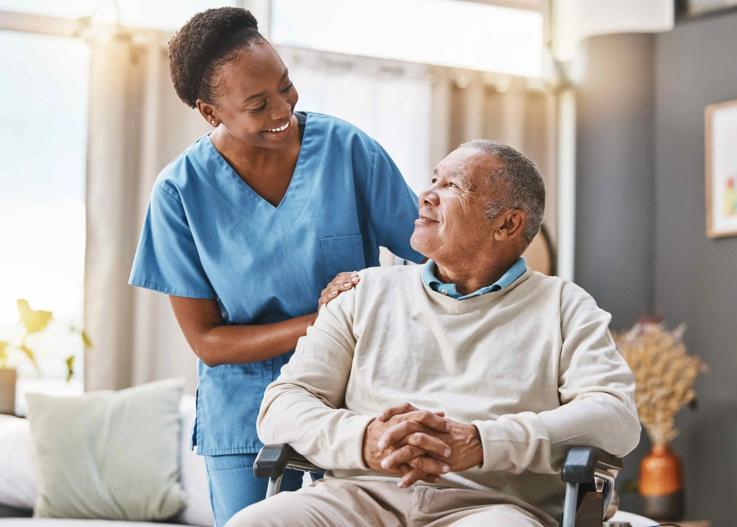 A nurse in blue scrubs smiling and talking to an elderly man in a wheelchair, who is smiling back, in a well-lit room.