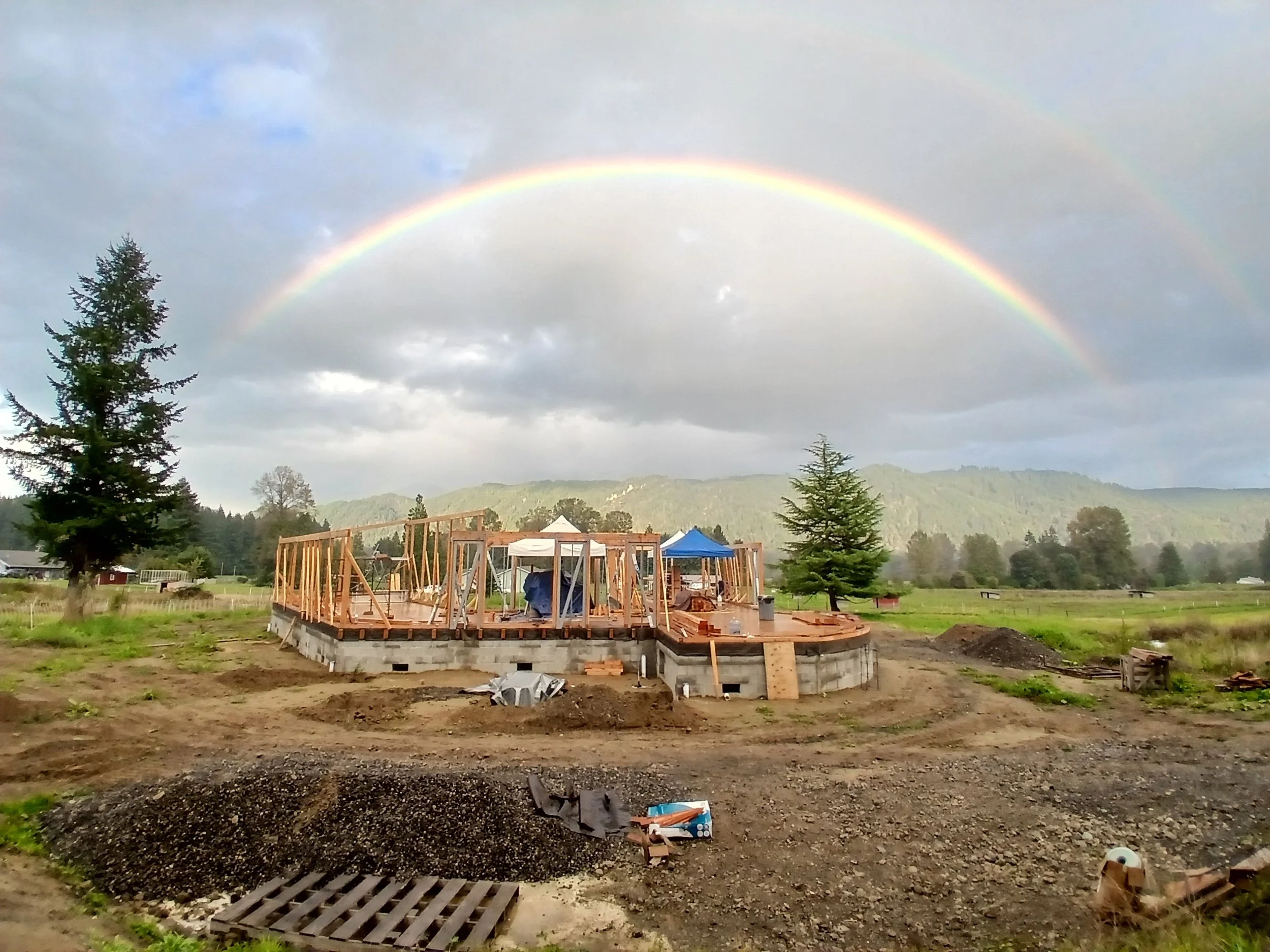 A strawbale home construction site with a wooden framework of a building under a double rainbow in a rural landscape.
